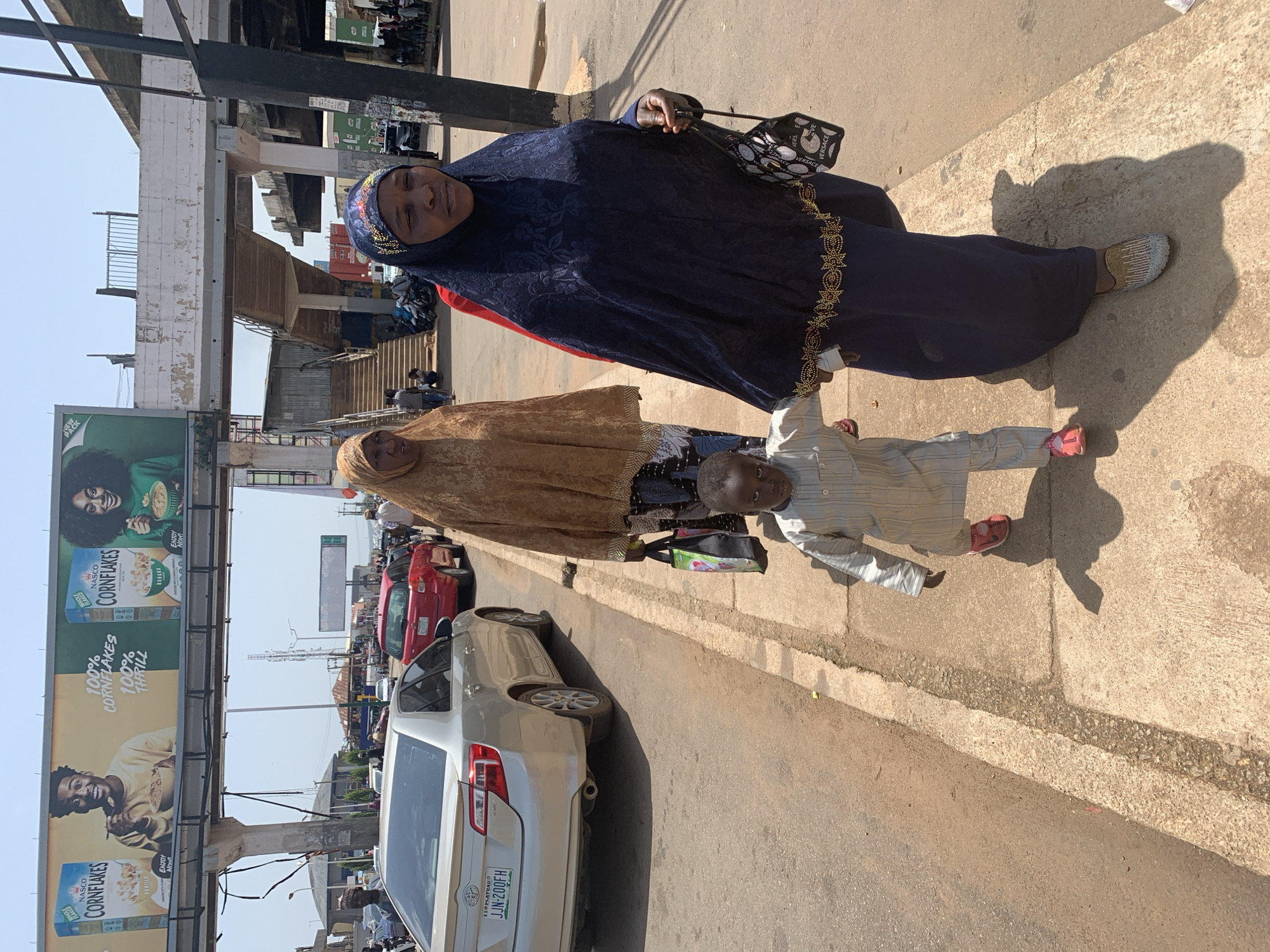 The image depicts a street scene in Jos, Nigeria, under a clear sky. In the foreground, a young boy in a light-colored striped outfit and pink shoes walks hand-in-hand with a woman wearing a dark blue hijab and matching long dress adorned with gold embroidery. Behind them, another woman in a tan hijab and patterned skirt walks, carrying a colorful shopping bag. To the left, a silver Toyota Camry is parked on the street, with its license plate showing "JJN-200FH". In the background, a pedestrian bridge with concrete supports and stairs is visible, along with billboards advertising "Nasco Cornflakes" featuring images of young adults. The environment appears to be an urban area with paved roads and sidewalks, and other vehicles and people are present in the distance, suggesting a bustling commercial or residential area.