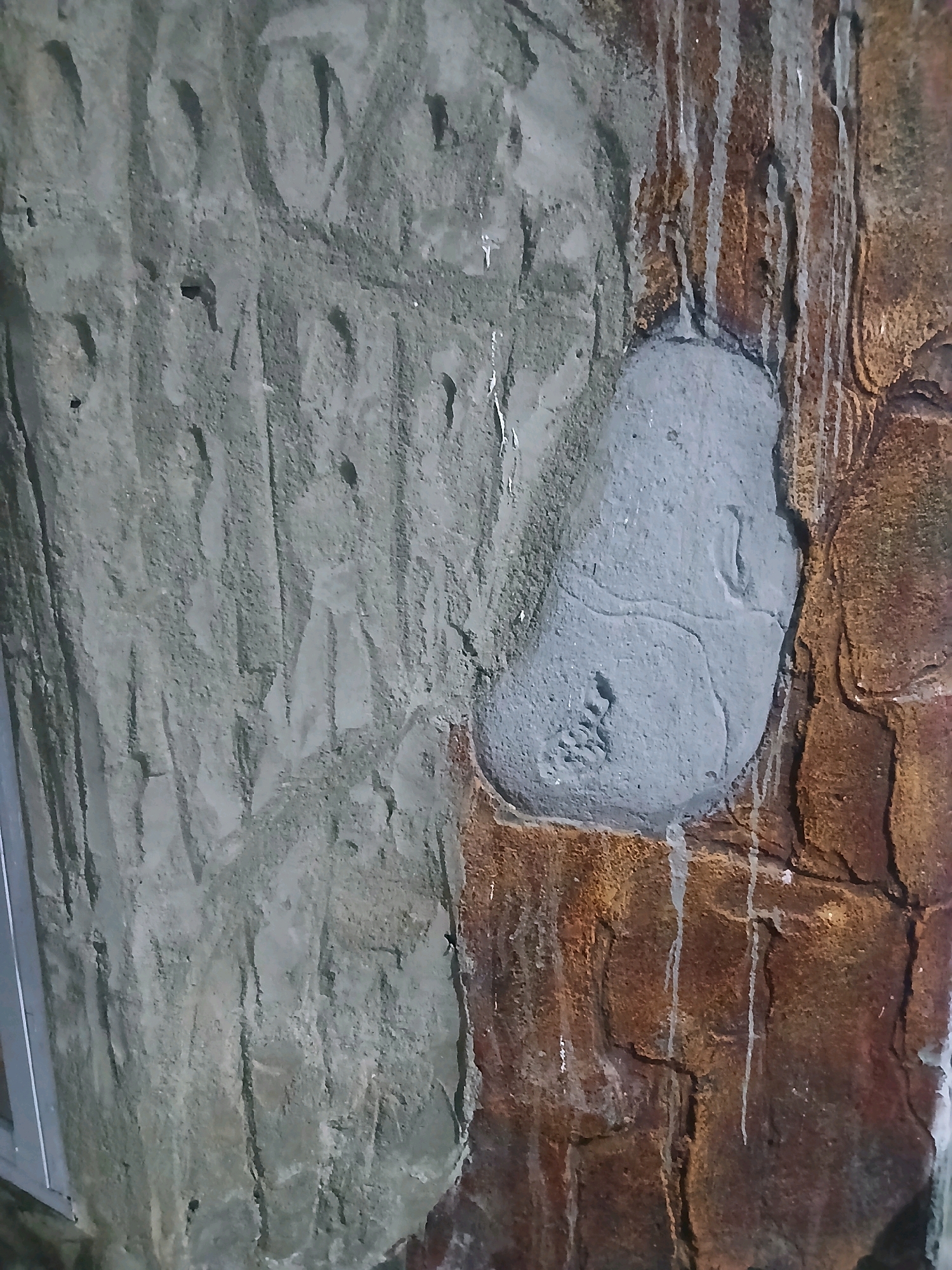 The image shows a close-up of a textured wall with two distinct sections. The left side of the wall is a rough, grey surface with vertical grooves and small indentations, possibly resembling unfinished concrete or a textured plaster. The right side of the wall features a darker, reddish-brown surface that appears to be made of simulated rock or stone. There are streaks of white, likely dried water or paint, running down this section. In the center of the image, a roughly oval-shaped patch of lighter grey material has been applied to the darker rock surface. This patch has a slightly raised, organic shape with faint lines or impressions within it, and a few small, clustered indentations at the bottom. No people or specific locations within Gorontalo, Indonesia, are identifiable in this image.