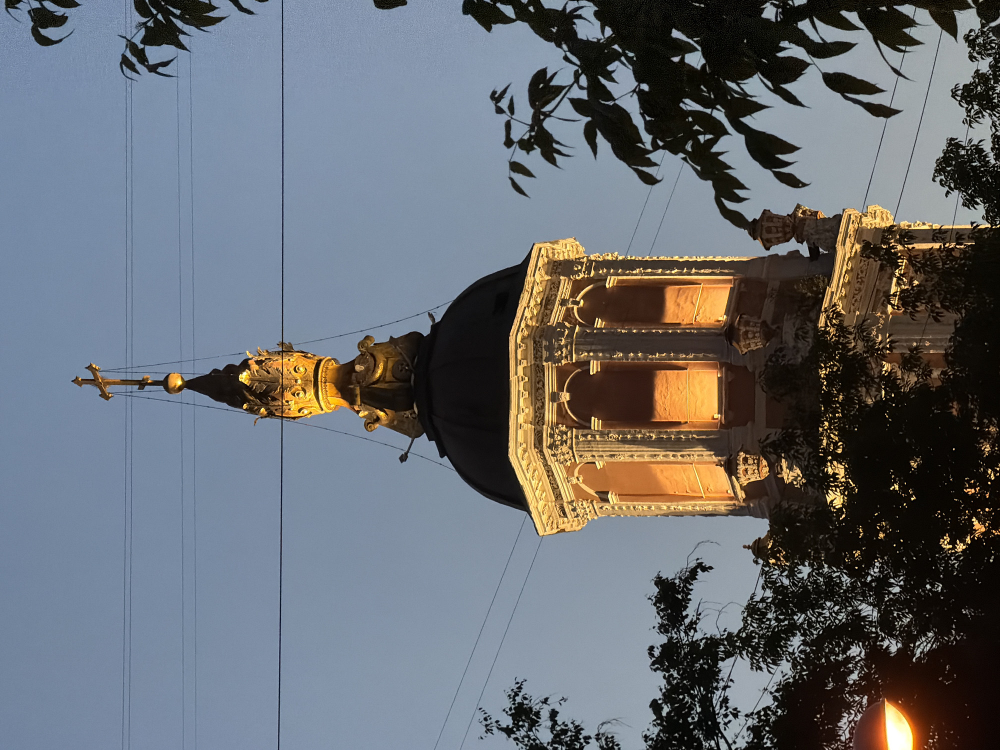 The image displays the upper section of the Menshikov Tower (Church of Archangel Gabriel) in Moscow, Russia, at twilight. The tower is illuminated with a warm, orange artificial light, starkly contrasting with the clear, cool blue twilight sky.
The tower's architecture features an ornate, multi-tiered structure. The visible lower section exhibits Baroque elements, including fluted pilasters, cornices, and arched niches that glow from internal illumination. Above this, a dark, rounded dome supports a gilded, crown-like ornament, from which a slender, golden spire ascends, topped by an Orthodox cross.
Several dark, thin utility wires are stretched horizontally across the sky, passing in front of the tower's upper sections. The foreground is occupied by the dark silhouettes of dense tree foliage, partially obscuring the lower portions of the tower on both the left and right sides of the frame. A glowing streetlamp, emitting warm light, is partially visible through the foliage in the lower-left corner. No individuals or specific activities are present in the scene.