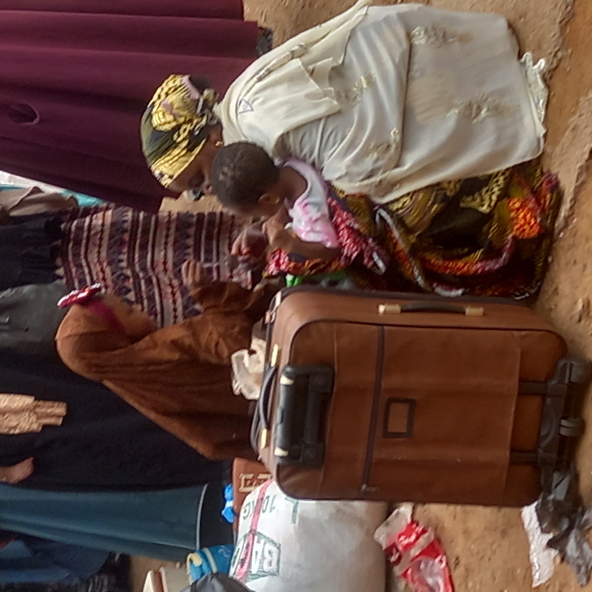 The image shows an indoor scene in Sardauna, Nigeria. An adult woman wearing a headscarf and light-colored top sits on the ground, attending to a small child in a light pink top. A suitcase rests beside them, and other bags and items are scattered on the floor.