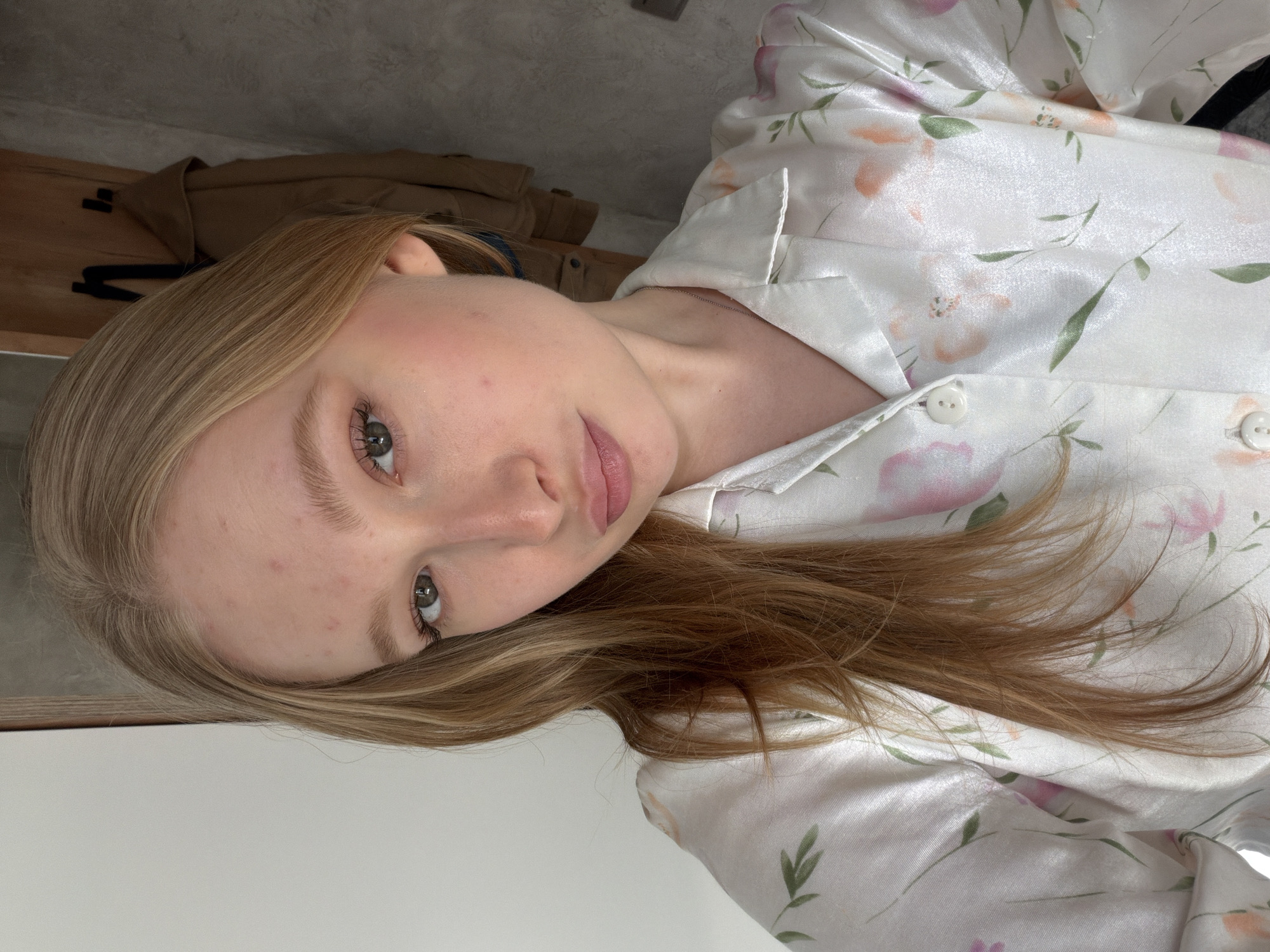 The image shows a person with fair skin, light brown and blonde hair, and light-colored eyes. The person is wearing a white satin or silk-like shirt with a floral pattern in shades of pink, orange, and green. The shirt is buttoned down, and the person is lying down. The background is blurry and includes what appears to be a wooden shelf with a brown jacket on it, and a white wall. The location is not discernable from the image, and there is no indication that the image was taken in Rossiyskiy, Russia.
