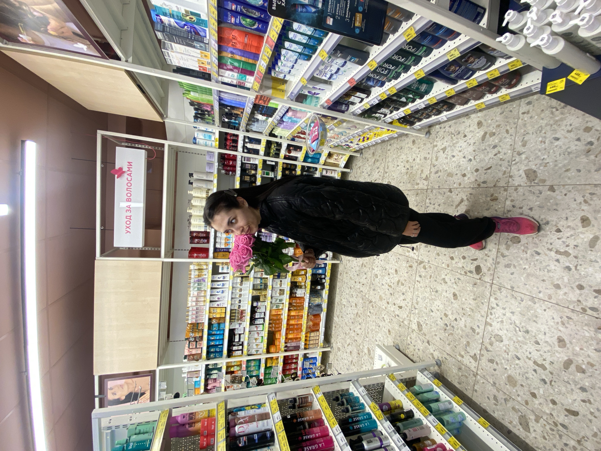 An indoor, portrait-oriented shot captures a woman standing in a supermarket or cosmetics store aisle, holding a bouquet of pink roses. The woman, with dark hair pulled back, is dressed in a dark, possibly quilted, jacket, dark pants, and bright pink sneakers. She looks towards the camera with a slight smile or smirk.
The setting is an aisle brimming with hair care products displayed on white shelves. Numerous shampoos, conditioners, and other beauty items in various colorful packages line the shelves, with some items bearing yellow price tags in Russian rubles (e.g., 469, 299, 239). Visible brands include "LA GRASE," "FOR," "Shauma," "DEONE," "LEX," "MON," "Dove," and "CLEAR CLEAR."
A prominent sign above the aisle clearly states "УХОД ЗА ВОЛОСАМИ," which translates to "HAIR CARE," designating the section. Other Russian text, such as "КУПИТЕ" (BUY) and numbers like "2" and "3," hint at ongoing promotions. The floor is made of light-colored terrazzo tiles, and the aisle is illuminated by fluorescent lights on the ceiling. The scene suggests a routine shopping excursion in Votkinsk, Russia, with the bouquet of flowers adding a personal touch, perhaps a gift or a fresh purchase.
