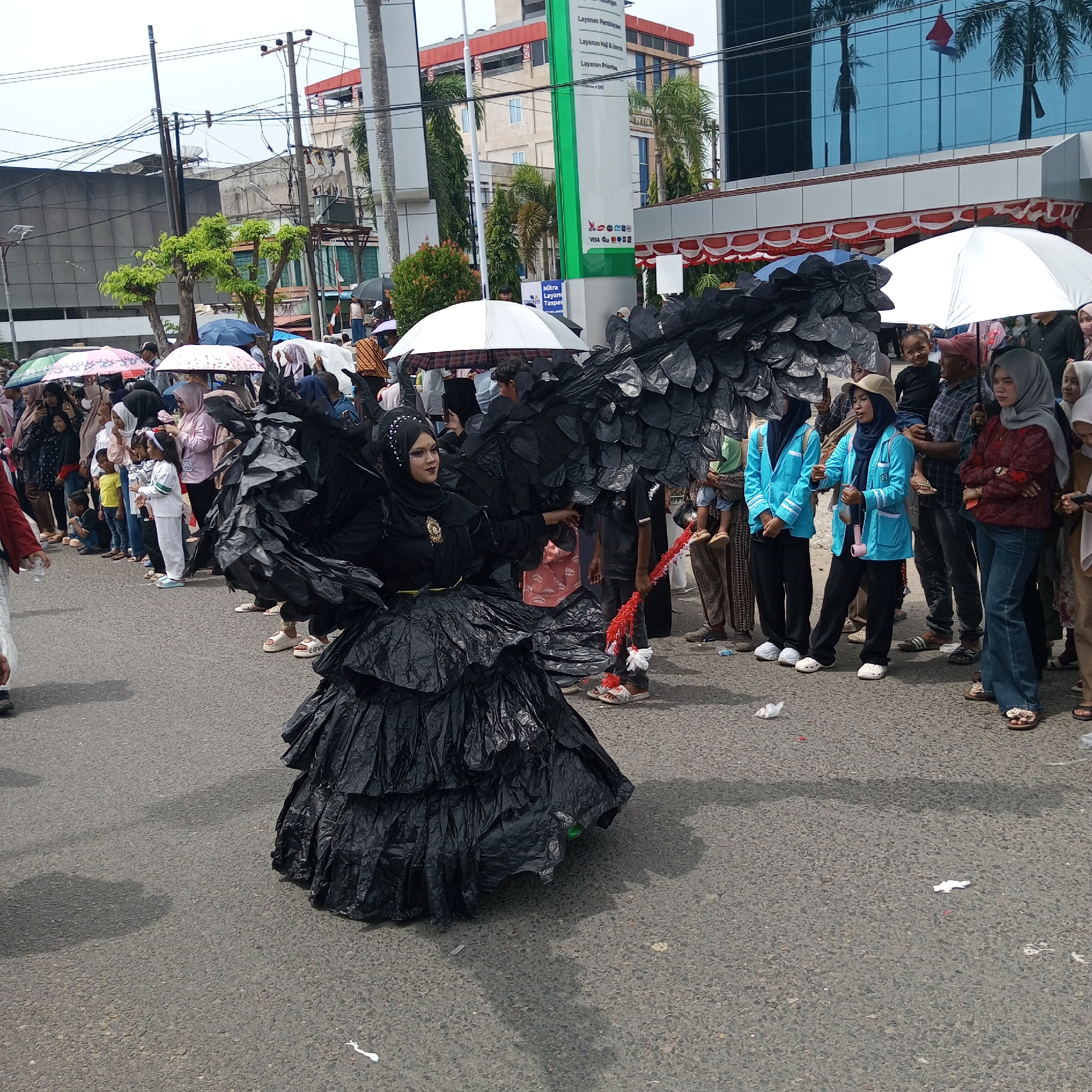 The image captures a street scene in Lhokseumawe, Indonesia, likely during an outdoor event or parade. In the foreground, a person in a dramatic black costume, resembling a large bird with exaggerated wings and a ruffled skirt made of what appears to be trash bags, is posing. The costume is dark and textured, with the skirt fanning out widely. The individual is wearing a hijab and makeup, suggesting a theatrical performance or a celebratory costume.
Behind the costumed figure, a crowd of spectators lines the street, many holding umbrellas, indicating overcast weather or a need for shade. The spectators are diverse, with people of various ages and genders. Some are looking towards the costumed individual, while others are engaged in conversation or watching something off-camera. Several individuals are dressed in casual clothing, while a group of people in bright blue jackets and black pants are standing together, possibly as participants or organizers of the event.
The background features urban elements, including buildings with modern architecture, one of which has a reflective blue glass facade. A tall green and white pillar with text and logos is visible, likely part of a public building or commercial establishment. Overhead, power lines and poles crisscross the sky, typical of an urban environment. The street itself is paved with asphalt, and there are scattered pieces of litter on the ground, including crumpled white paper. The overall atmosphere appears to be that of a community gathering or cultural event taking place in a city.