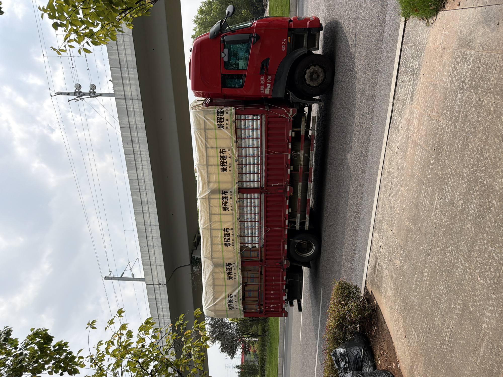 A red flatbed truck is driving on a road under an overpass, carrying rolls of material covered by a white tarp. The truck has Chinese writing on its side, including "景程篷布" which translates to "Jingcheng Tarpaulin." The overpass appears to be a train track or elevated highway with power lines visible. The scene is likely in Jiangjin, China, given the writing on the truck. The truck is in motion, indicated by the slight blur and the perspective. There are no people clearly visible in the image. The environment is an urban or suburban setting with trees and a cloudy sky. The road is asphalt, and there is a sidewalk with some black trash bags. The overall impression is a snapshot of daily transportation and infrastructure in a Chinese city.