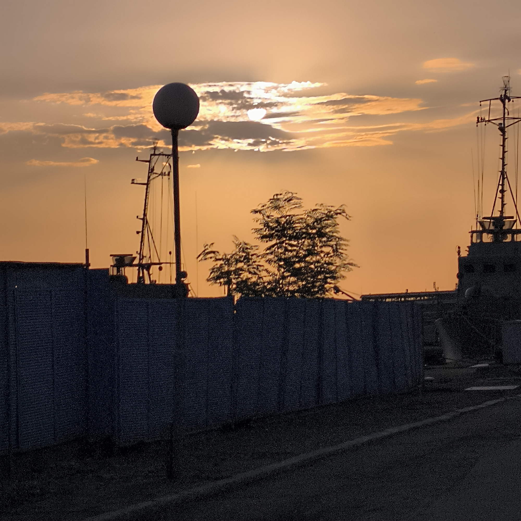 The photograph captures a sunset over a waterfront area in Varna, Bulgaria. The sky is dominated by warm orange and yellow hues, with clouds partially obscuring the setting sun, creating rays of light. In the foreground, a dark, textured fence runs horizontally across the image, with a dark road or walkway in front of it. To the left, silhouetted against the sky, is a tall pole topped with a spherical object, likely a light or sensor. Behind the fence, the masts and superstructures of ships or industrial equipment are visible in silhouette. A cluster of trees with feathery foliage is also present, silhouetted against the sky. The overall impression is one of an industrial or port setting at dusk.