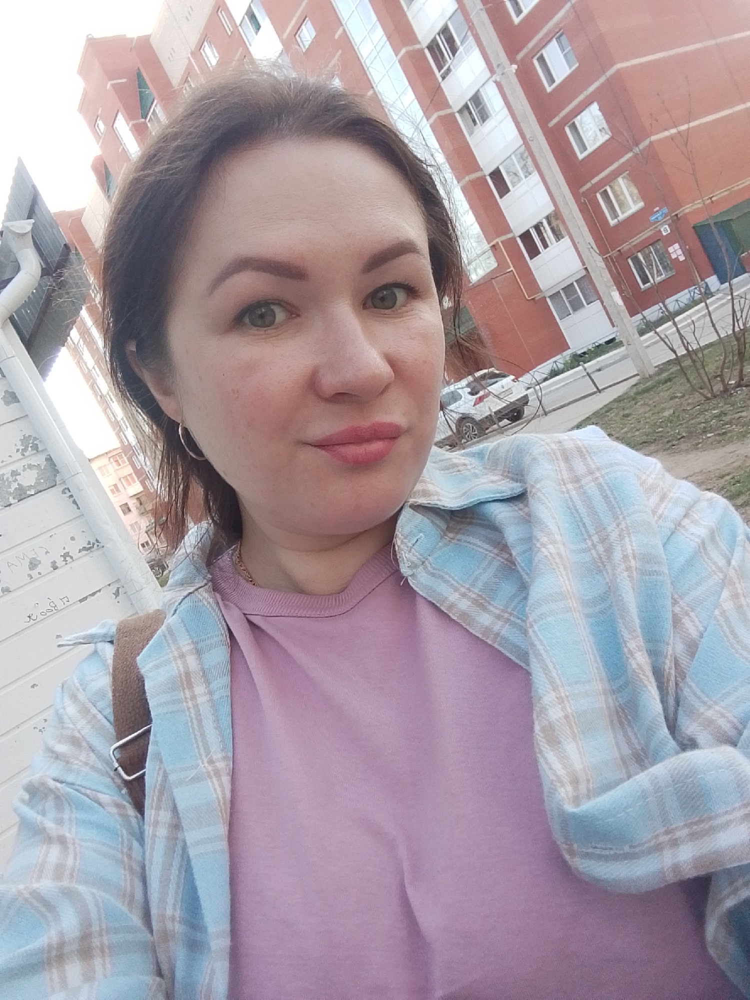A woman with fair skin, dark hair pulled back, and wearing a pink t-shirt and a blue and white plaid shirt is taking a selfie. She has light green eyes and pink lipstick. She is wearing hoop earrings and a thin gold necklace. Behind her is a red brick building with white-framed windows, a white pole, and some trees. A white car is visible in the background. The scene is located in the city of Chaikovsky, Russia. A white gutter and a section of a white-painted wall are visible to the left.