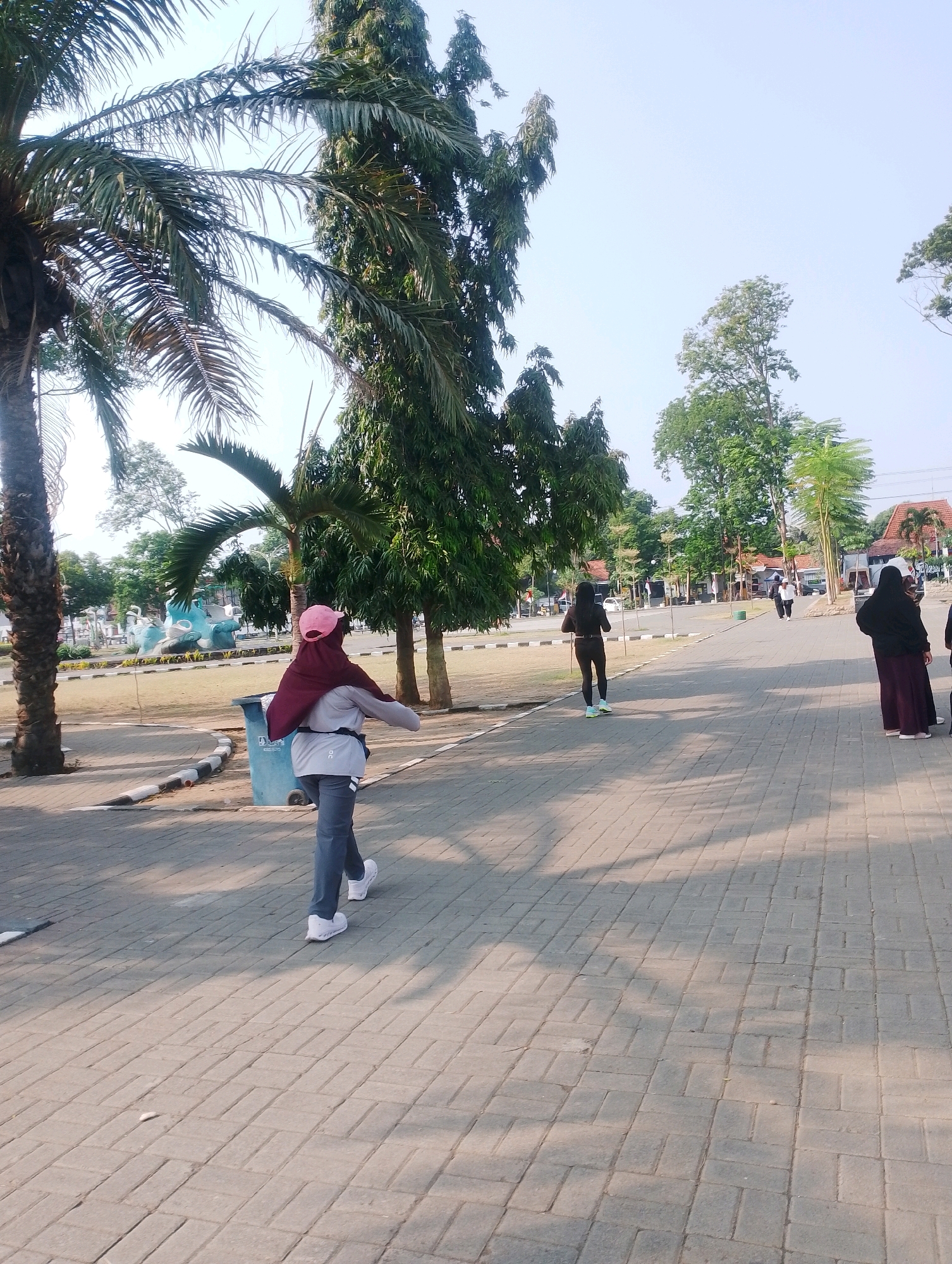 The image shows a paved outdoor area with several people walking. In the foreground, a person wearing a maroon hijab, grey shirt, and grey pants is walking away from the camera. A pink baseball cap is visible under the hijab. To the right, a person in a black long-sleeved top and black leggings walks in the same direction. Further in the background, other individuals are walking, including a person in a dark purple abaya and headscarf. The environment features palm trees on the left and a variety of other trees and buildings in the distance. A blue trash can is visible next to the person in the foreground. The ground is paved with interlocking brick pavers. The image appears to be taken in a public park or walkway. This scene is likely located in Probolinggo, Indonesia, a city known for its coastal location and tropical climate, which would explain the presence of palm trees. The bright sunlight suggests it is daytime.