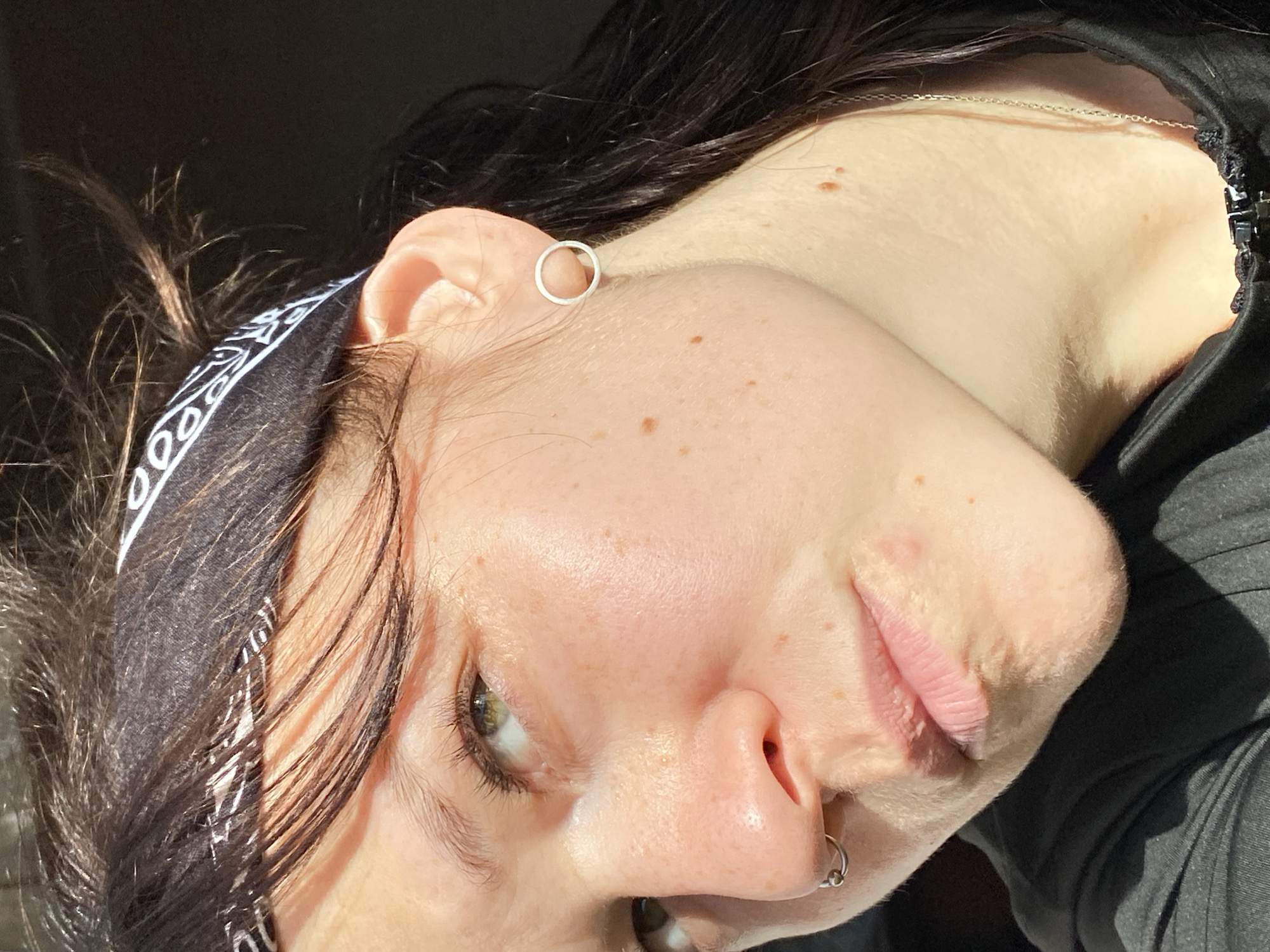 A close-up, angled portrait of a person with fair skin. The person has dark hair partially covered by a black bandana with white designs. They have a circular earring in their left ear and a ring piercing in their left nostril. The person has light green eyes. The person is wearing a dark top and a silver necklace. The image location cannot be identified.