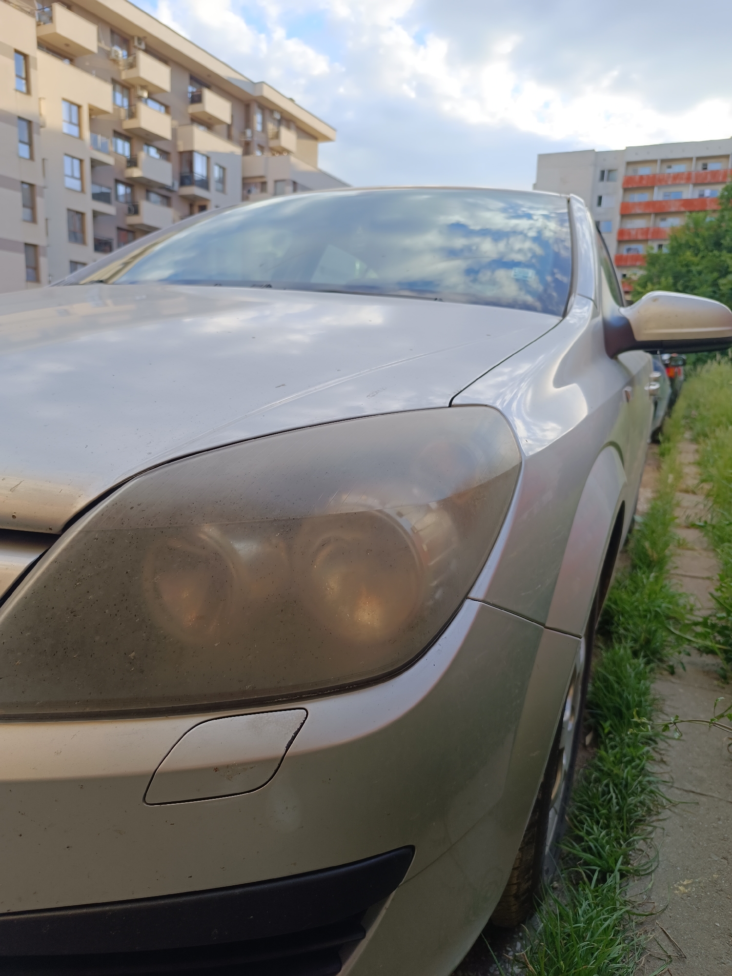 The image depicts a silver Opel Astra parked on a concrete curb bordered by overgrown grass. The car's headlight is heavily oxidized and dirty. In the background, there are residential buildings, one appearing newer with beige facade and balconies, and another older building with red-trimmed balconies. Based on the architectural style and vegetation, this image could possibly be in Sofia, Bulgaria.