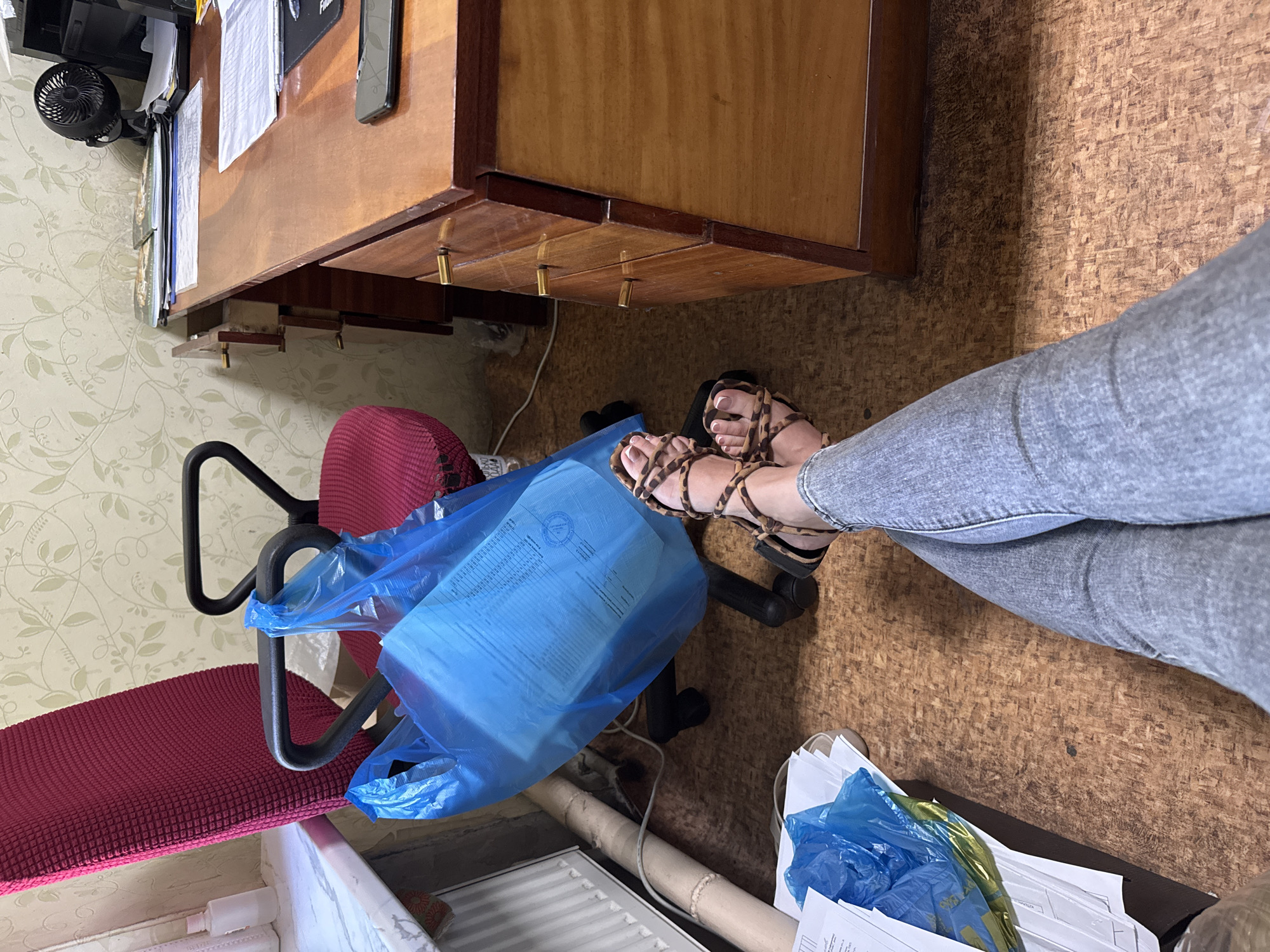 A person's legs and feet, clad in light-wash denim jeans and leopard-print sandals, are positioned in the foreground, crossed at the ankles. The person is seated in an office environment. To the left of the frame, a bright pink office chair is visible, with a large blue plastic bag hanging from its armrest. The bag appears to contain papers or documents. Behind the chair, a wooden desk with brass drawer pulls is situated. On the desk, a computer monitor, a small black fan, and various papers are arranged. The floor is covered with a cork-like material, and a white radiator is present along the wall to the left. The background features wallpaper with a floral pattern. The lighting suggests an indoor setting during the daytime.