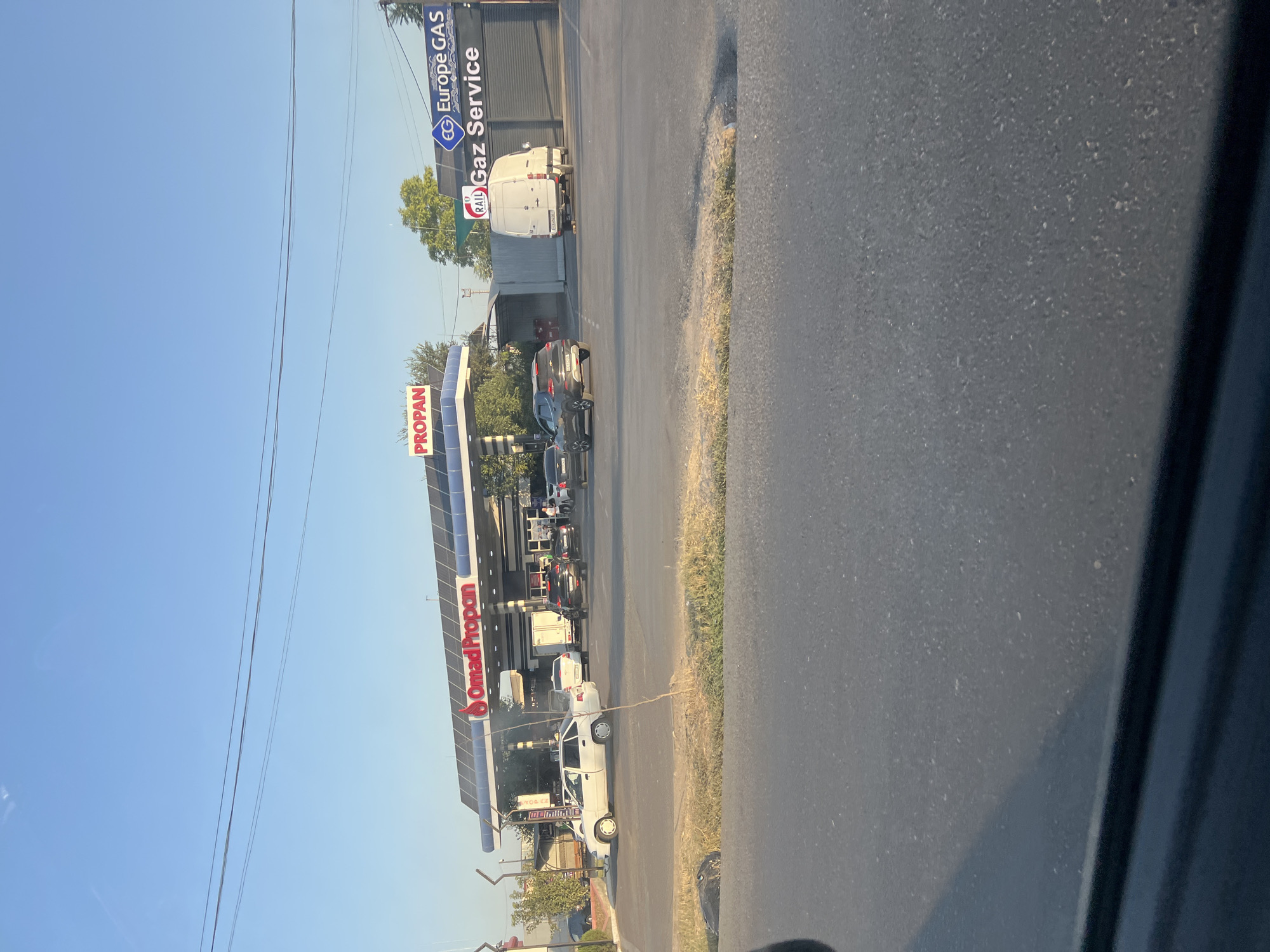 A gas station named "Omaq Propan" with a "Propan" sign is visible under a bright blue sky. To the right, a sign for "Europe GAS Gaz Service" with an "EG" logo is also present. Several cars are at the gas station, including a white sedan, a white van, and other darker colored vehicles. The ground appears to be asphalt. The image was taken in Erkin, Uzbekistan.