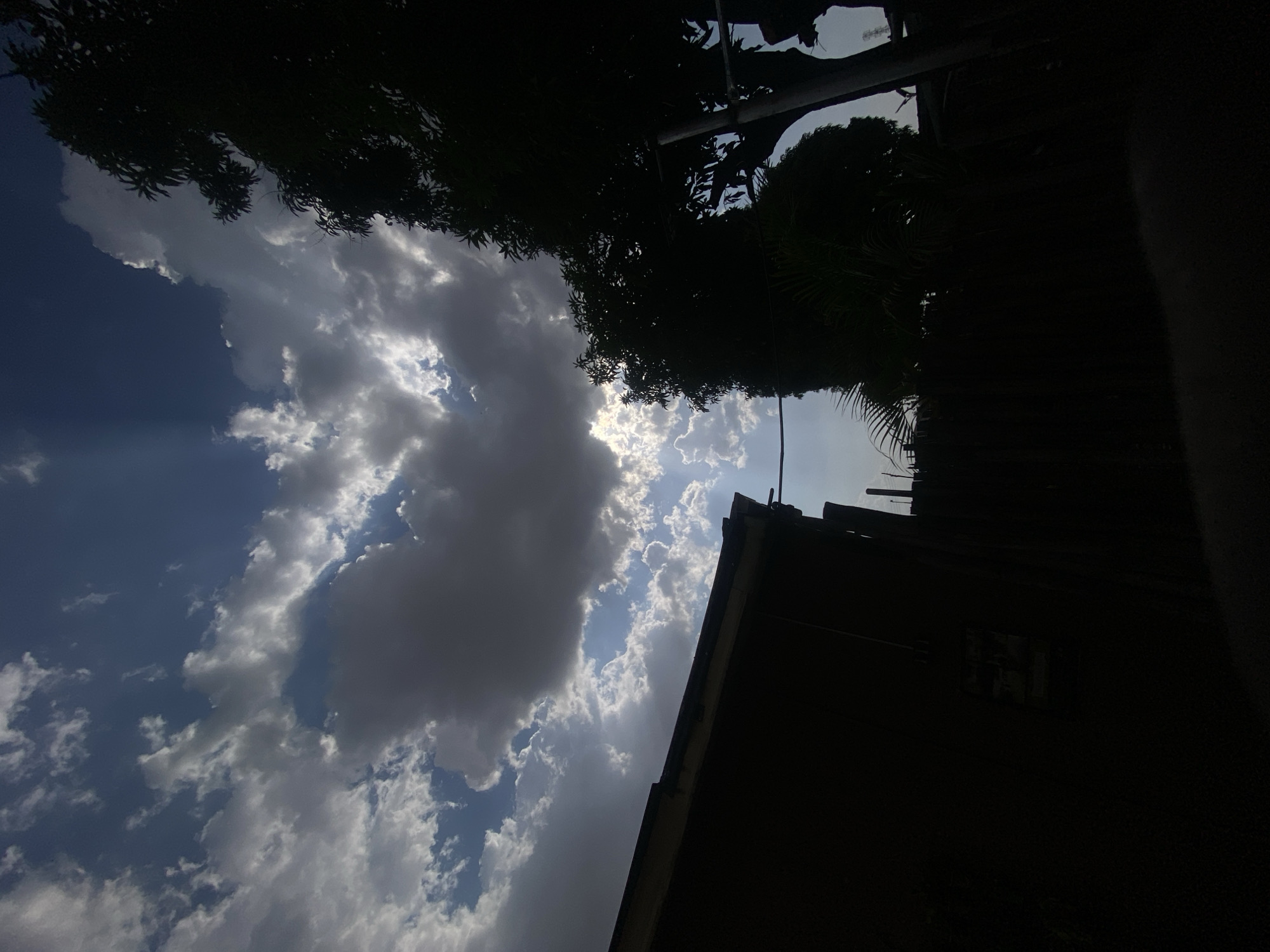 This image depicts a partially obscured sky in Barden Yana, Nigeria. Cumulus clouds of varying density fill the left side of the frame, with patches of blue sky visible between them. Sunlight filters through the clouds. Above, silhouetted tree foliage and branches occupy the top edge of the image. A dark building with a rectangular window and angular roof lines is partially visible on the right side, obscuring a large portion of the sky. The scene suggests a combination of natural elements and urban structures in Barden Yana.