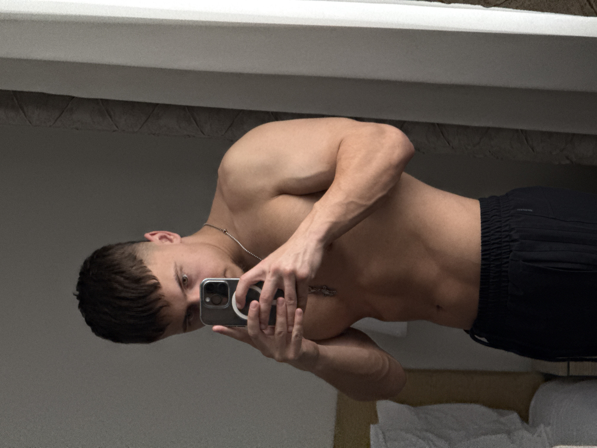 A male individual is depicted in a mirror selfie, shirtless and wearing black athletic shorts with an elasticated, ribbed waistband. He possesses a muscular build, visible in his torso and arms, and has short, dark hair with a distinct fringe and faded sides. A silver-colored chain with a pendant hangs around his neck. The subject holds a dark-colored smartphone, featuring a circular magnetic accessory on its back, positioning it to capture his reflection. His gaze is directed towards the smartphone's camera. The indoor background consists of a light-colored panel or headboard on the left, an adjacent wall with a dark, textured, possibly geometric pattern, and a vertical white structural element on the far right. The scene is situated in an indoor setting, specified as being within the city of Vozdovac Urban Municipality, Serbia.