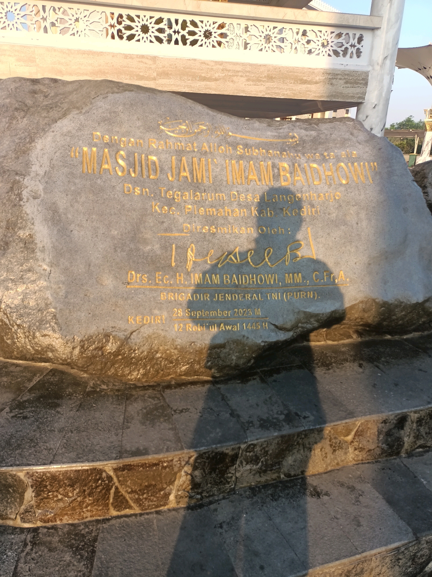 The image shows a stone plaque with golden inscriptions, located at the entrance of a mosque in Plemahan, Indonesia. The inscription reads "MASJID JAMI IMAM BAIDHOWI" and provides details about its location: Dsn. Tegalarum Desa Langenharjo, Kec. Plemahan Kab. Kediri. The plaque also mentions that it was inaugurated by Drs. Ec. H. IMAM BAIDHOWI, MM., C.Fr.,A., BRIGADIR JENDERAL TNI (PURN.) on September 28, 2023 M, corresponding to 12 Rabi'ul Awal 1445 H. A shadow of a person is visible on the plaque, cast by the person taking the photograph. The stone plaque is placed on a tiered stone structure, possibly steps leading up to the mosque. The background shows part of the mosque's architecture with intricate patterns and a portion of the sky.