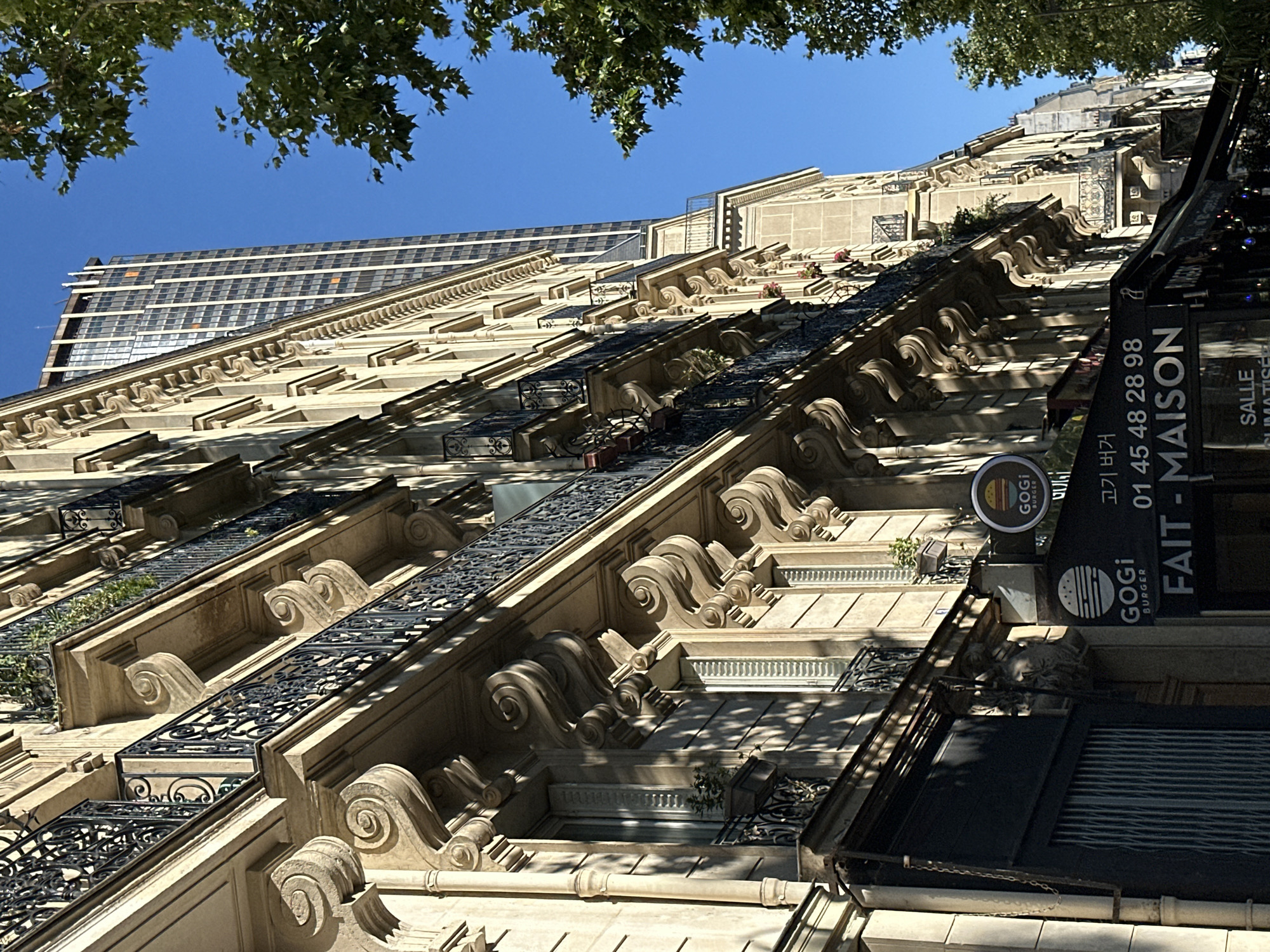 This is a street-level, eye-level, medium shot capturing a section of a Haussmannian building facade in Paris, France. The building, characterized by its ornate balconies with wrought-iron railings and decorative stone carvings, is bathed in sunlight, casting strong shadows. Several windows with closed shutters are visible. Below the building's facade, a dark awning is extended, featuring signage for "GOGi BURGER" and the French phrase "FAIT - MAISON". A phone number, "01 45 48 28 98", is also displayed on the awning. A modern, tall skyscraper with a glass facade is visible in the background, contrasting with the classical architecture of the foreground building. Lush green tree foliage is present in the upper right quadrant of the image, and the sky is a clear, bright blue. The scene appears to be devoid of people.