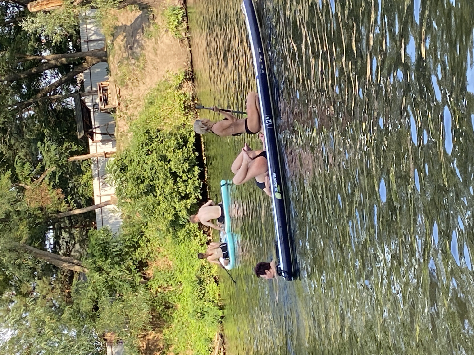 In Petrivka, Ukraine, on a calm body of water, four individuals are engaged in recreational activities. In the foreground, a person is partially submerged in the water, with only their head and shoulders visible, and a dark blue inflatable raft beside them. Further back, two individuals are on light blue paddleboards. One, a male with his back to the camera, is seated. The other, a female facing away, is also seated and holds a paddle. To the right, a female on a long, dark blue paddleboard is seated, holding a paddle and looking to her left. The background features lush green foliage, trees, a fence, and a wooden structure with a roof and seating. The water reflects the sky and surrounding greenery.