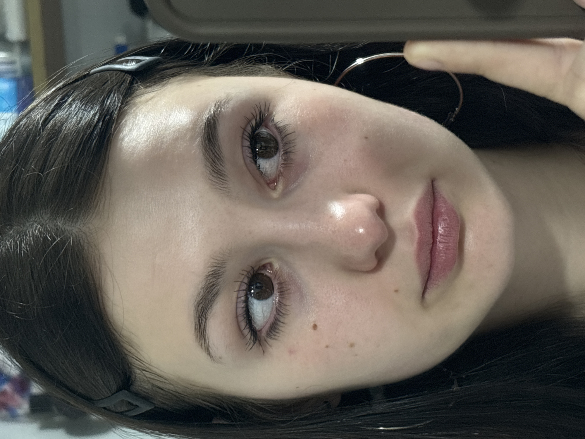 A young Caucasian woman with fair skin, dark brown hair, and brown eyes is taking a selfie. She has a small mole on her left cheek and another near her mouth. She is wearing a silver hoop earring and a dark hair clip is visible in her hair. The background is blurred and indistinct, but appears to be an indoor setting. There is no information available to determine if the location is Фоки, Russia.