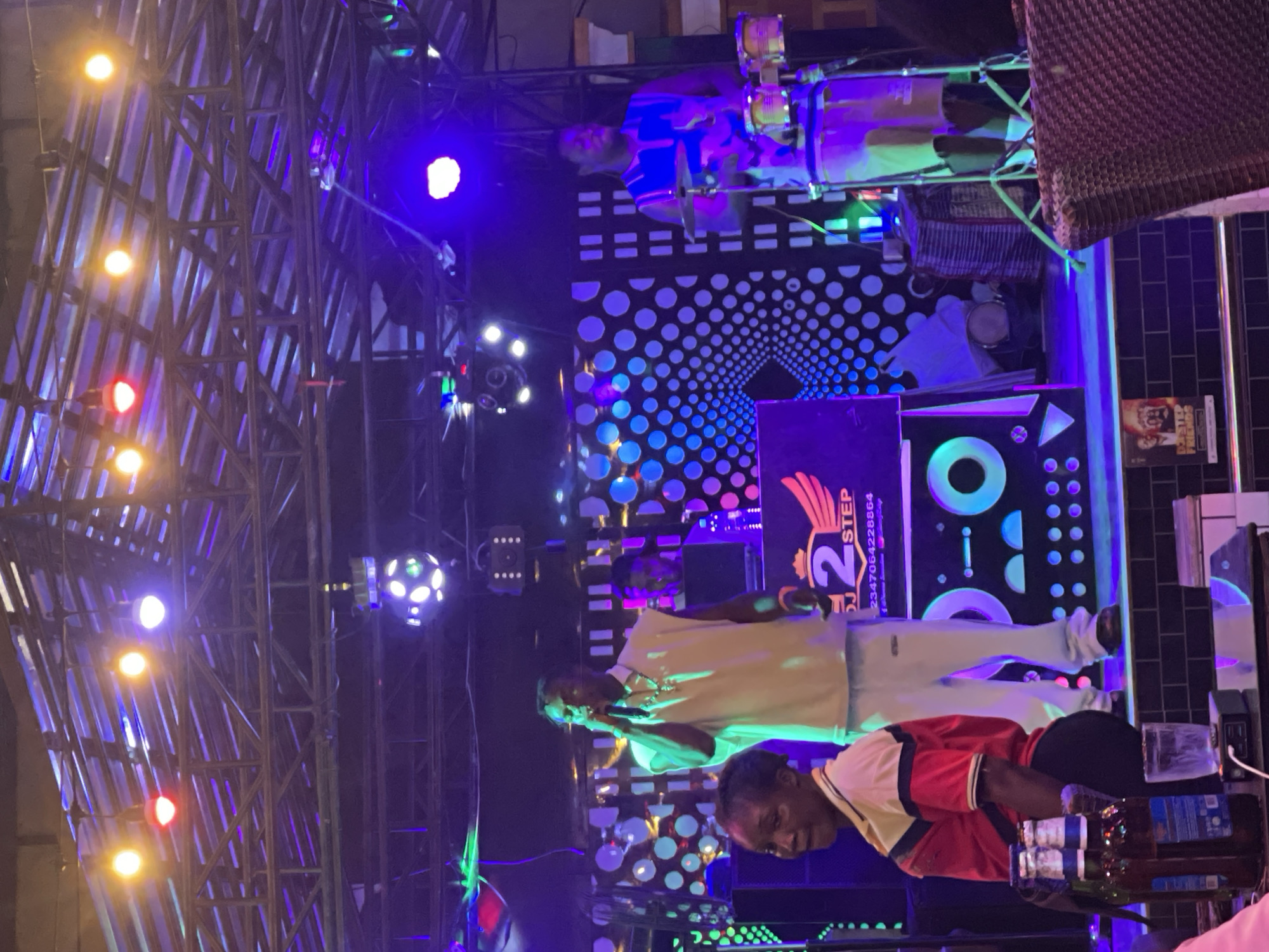 The scene depicts a live musical performance at a venue in Obosi, Nigeria. A male lead vocalist, dressed in a light-colored sleeveless shirt and light pants, stands center stage, holding a microphone and singing. Behind him, a male DJ operates a mixing console and laptop. To the right, a male percussionist, wearing a blue and white striped sleeveless shirt and light shorts, plays a set of small drums and a cymbal. The stage features a backdrop with circular and vertical bar patterns, illuminated by vibrant blue and purple lighting. An overhead metal truss structure supports multiple stage lights, including red, yellow, and white spotlights, as well as a disco ball. The DJ booth bears a "2 STEP DJ" logo with a phone number. In the foreground on the left, a woman with dark skin, wearing a red, yellow, and white polo shirt, stands beside a table. On this table are two brown beer bottles, a clear glass, and a small electronic device with a white cable. The stage is raised, with its base displaying a brick pattern and a "DOORSTEP FRIENDS" promotional poster. A brown wicker chair is partially visible in the far right foreground, suggesting a seating area for patrons. The overall environment is dark, dominated by the colorful illumination from the stage lights.