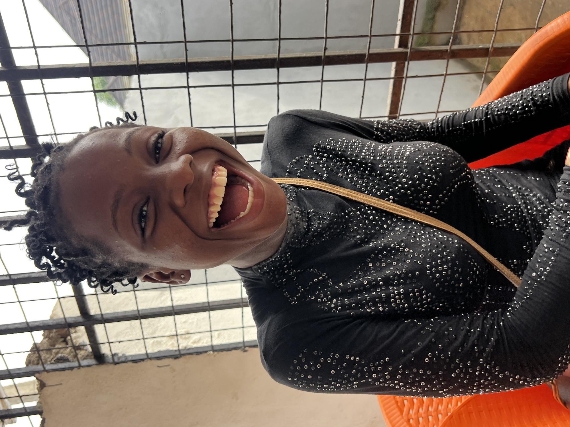 A medium shot portrait shows a Black woman with dark skin and curly hair, wearing a black long-sleeved shirt with a high neckline adorned with silver sequins. She is seated in an orange plastic chair, with her head tilted back and her mouth wide open in a laugh. A thin, light brown strap is visible crossing her chest. She has a small stud earring in her visible ear. Behind her, a large window with a grid of metal bars is present. The background shows some of the interior of a building with a textured wall and a glimpse of an outdoor environment with a roofline and greenery. The scene appears to be located in Nkpor, Nigeria.