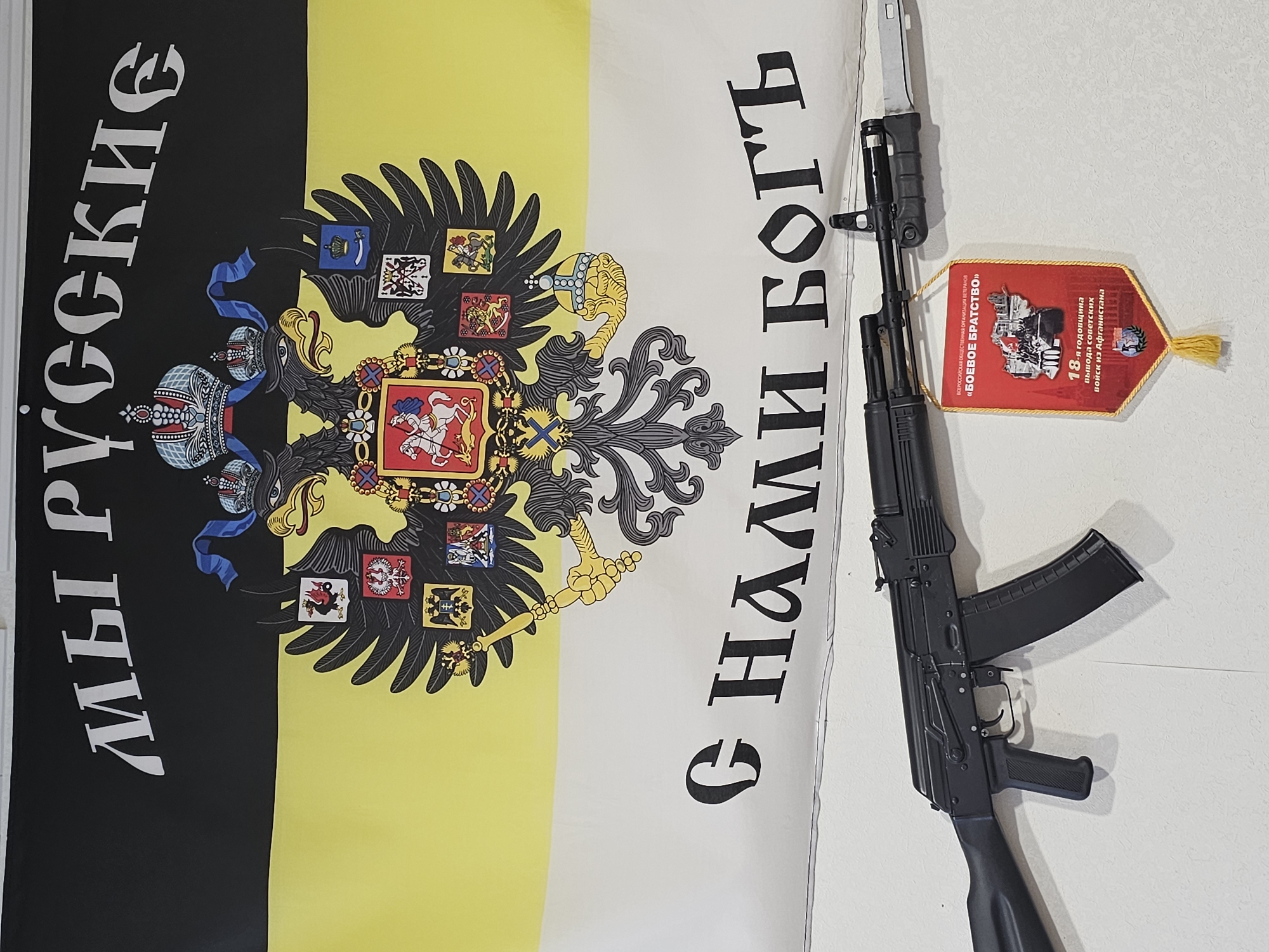 The image depicts a vertically oriented tricolor flag with black, yellow, and white stripes, bearing a central emblem of a double-headed eagle with various coats of arms. To the left of the flag, vertical Cyrillic text reads "МЫ РУССКИЕ" (WE ARE RUSSIANS), and to the right, also in Cyrillic, "С НАМИ БОГЪ" (GOD IS WITH US). Superimposed on the right side of the image is a black AK-pattern rifle with a bayonet attached. Next to the rifle, there is a red pennant with gold fringe, featuring Cyrillic text and imagery referencing the "BOEBOE BPATCTBO" (COMBAT BROTHERHOOD), an organization of veterans, and the 18th anniversary of the withdrawal of Soviet troops from Afghanistan. The environment appears to be an interior setting, with a plain white wall in the background. There is no explicit indication that this image was taken in Moscow, Russia.