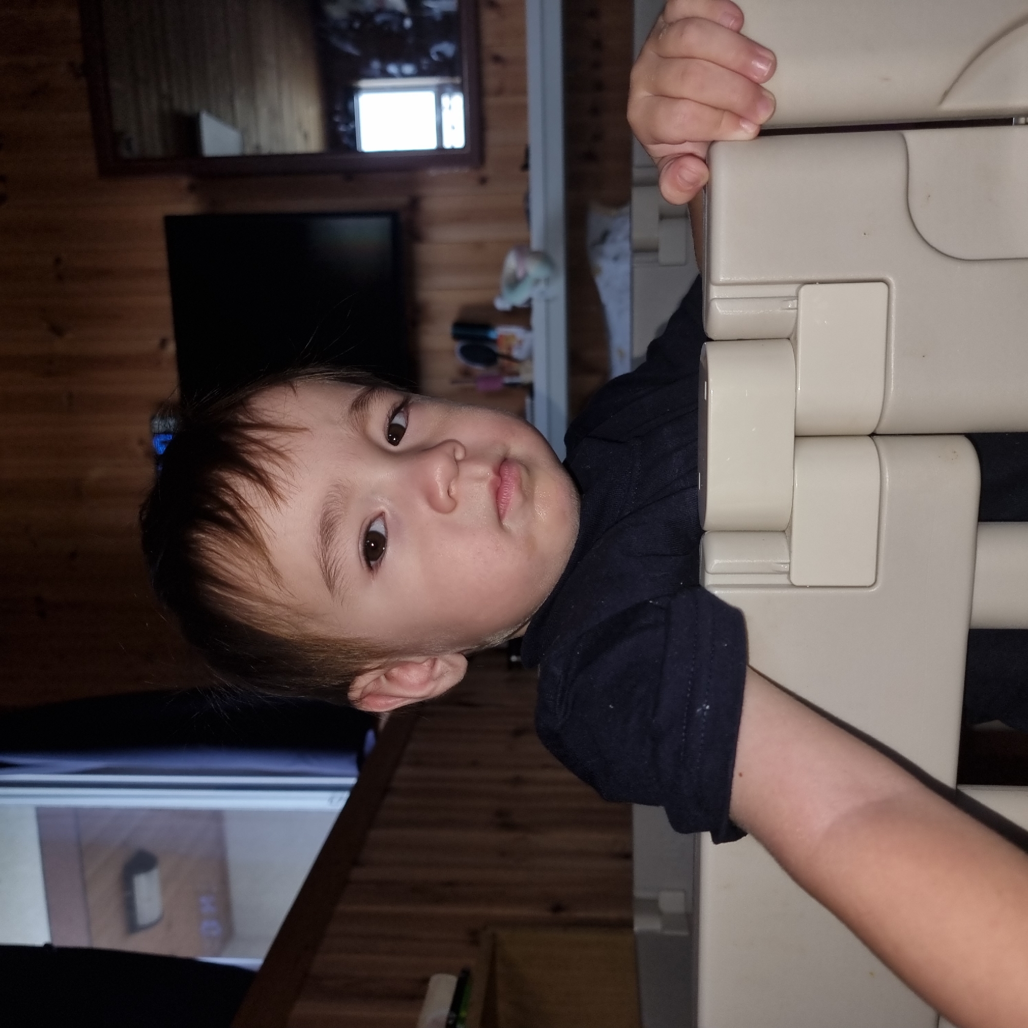 A young child, appearing to be of toddler age, is positioned inside a cream-colored playpen. The child has light brown hair, dark eyes, and a neutral expression. They are wearing a dark t-shirt. The background consists of wood-paneled walls, a television screen, and a mirror reflecting a window. The overall lighting suggests an indoor evening setting. The image does not contain enough information to determine the specific location within Dunnae-myeon, South Korea.