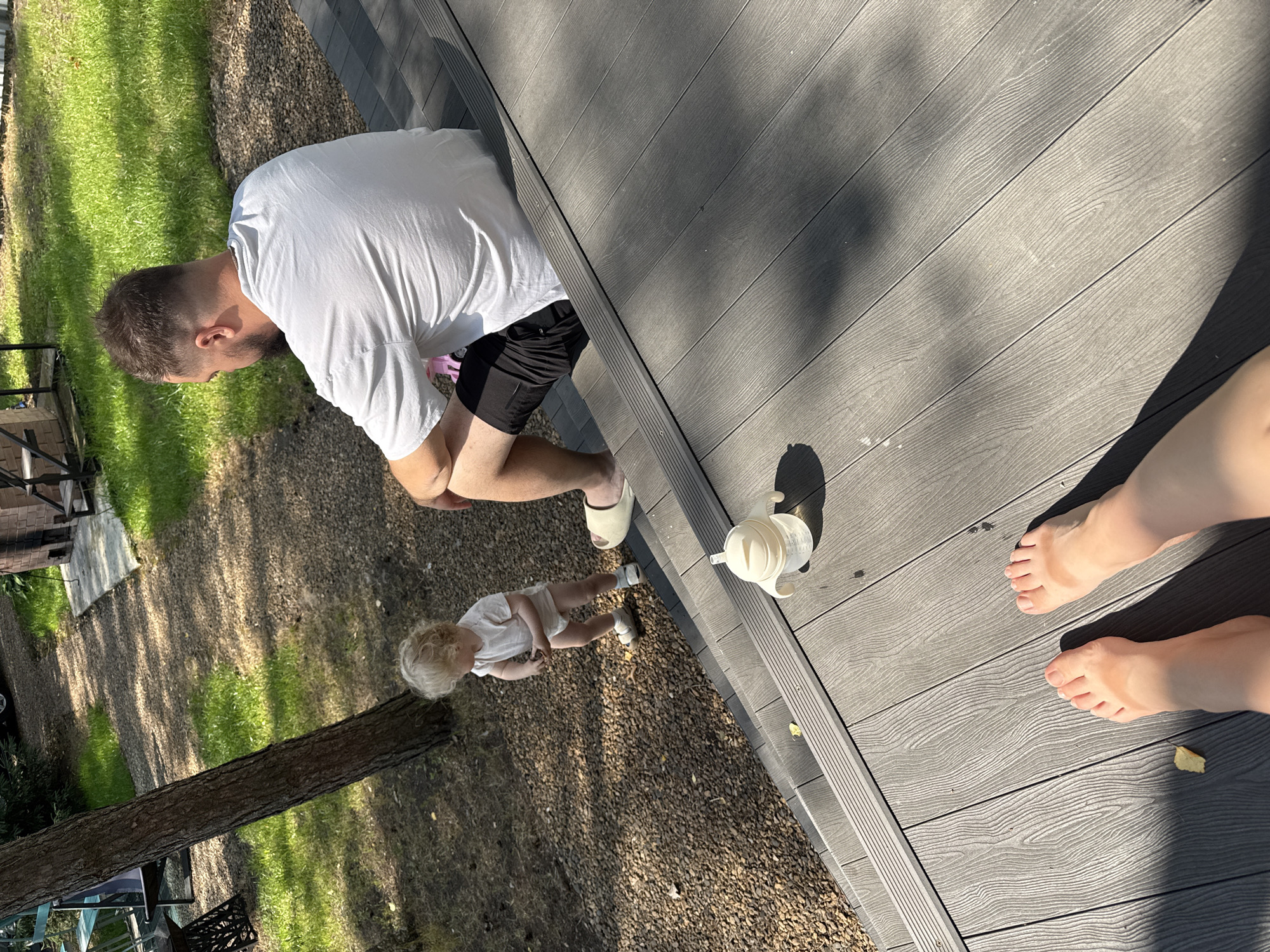 In Prokudino, Russia, on a sunny day, a toddler with blond curly hair, wearing a light-colored onesie and white shoes, stands on a gravel path next to a tree. A man in a white t-shirt and black shorts is seated on a wooden deck. A sippy cup is placed on the deck. In the foreground, two bare feet are visible on the wooden deck, with visible tan lines. The background shows a grassy area and a brick structure.
