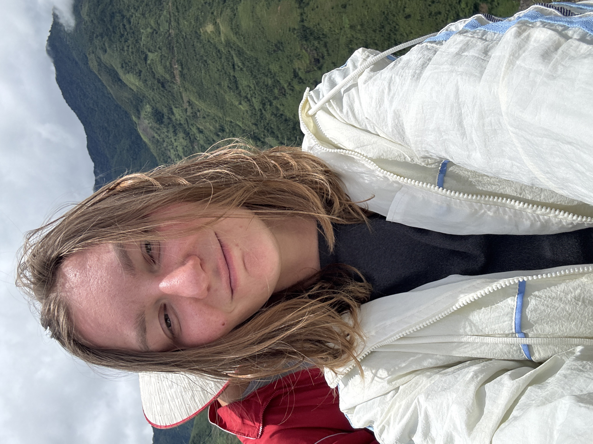 A young person with blonde hair pulled back is in the foreground, taking a selfie. They are wearing a white jacket with blue trim and a dark t-shirt underneath. Partially visible to their left is another person in a red jacket and a conical hat. In the background, lush green mountains rise under a cloudy sky. This scene appears to be captured in the mountainous terrain surrounding Bắc Yên, Vietnam.