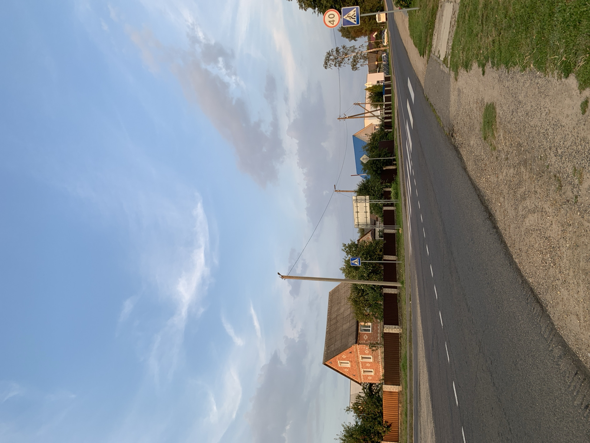 The image shows a street in the city of Kapyĺ, Belarus. A two-story brick house with a grey roof is visible on the left side of the street, behind a brown fence. A pedestrian crossing is marked on the asphalt road in the foreground. On the right side of the road, there are traffic signs indicating a pedestrian crossing and a speed limit of 40 kilometers per hour. A white bus is seen in the distance. The sky is partly cloudy.