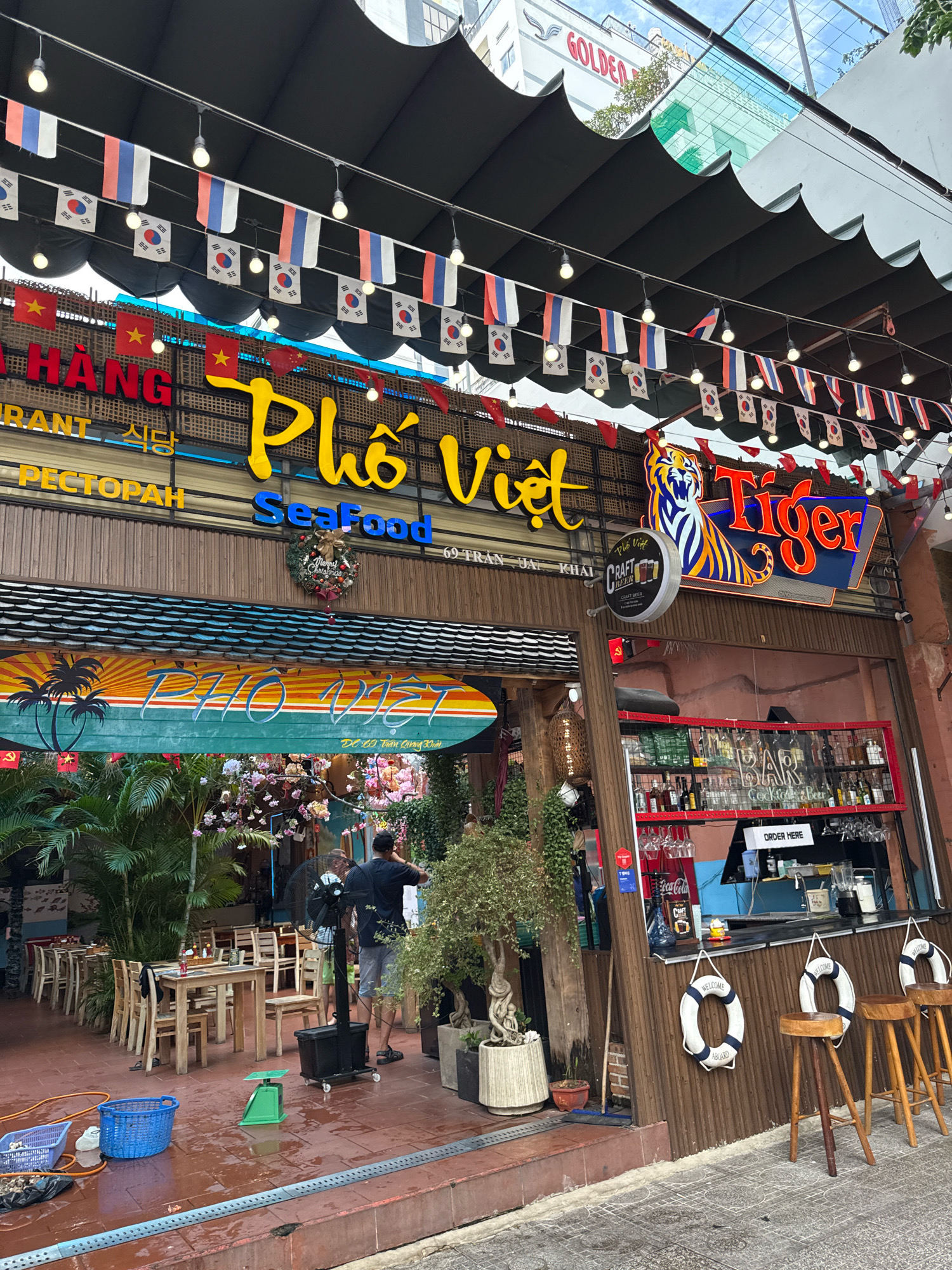 The image captures the exterior of a restaurant named "Pho Viet" located at 69 Tran Quang Khai Street in Nha Trang, Vietnam. The restaurant has a prominent yellow sign with Vietnamese characters and the word "Seafood" in blue. Above the entrance, a large neon sign featuring a tiger and the word "Tiger" is illuminated. Flags from various countries, including South Korea, Russia, and Thailand, are strung across the front of the restaurant, along with red triangular flags.
The outdoor seating area is visible, with tables and chairs set up on a tiled patio. Lush green plants and flowering trees surround the seating area, creating a vibrant atmosphere. A bar area is set up inside the restaurant, with bottles of alcohol and glasses on display. A "Welcome Aboard" lifebuoy decoration hangs near the bar.
Two individuals are visible in the image: one person is standing near a large black fan, and another is seated at an outdoor table. The overall scene suggests a bustling and lively restaurant, popular with both locals and tourists, given the international flags displayed. The architectural style and the presence of palm trees hint at a tropical coastal setting, consistent with Nha Trang.