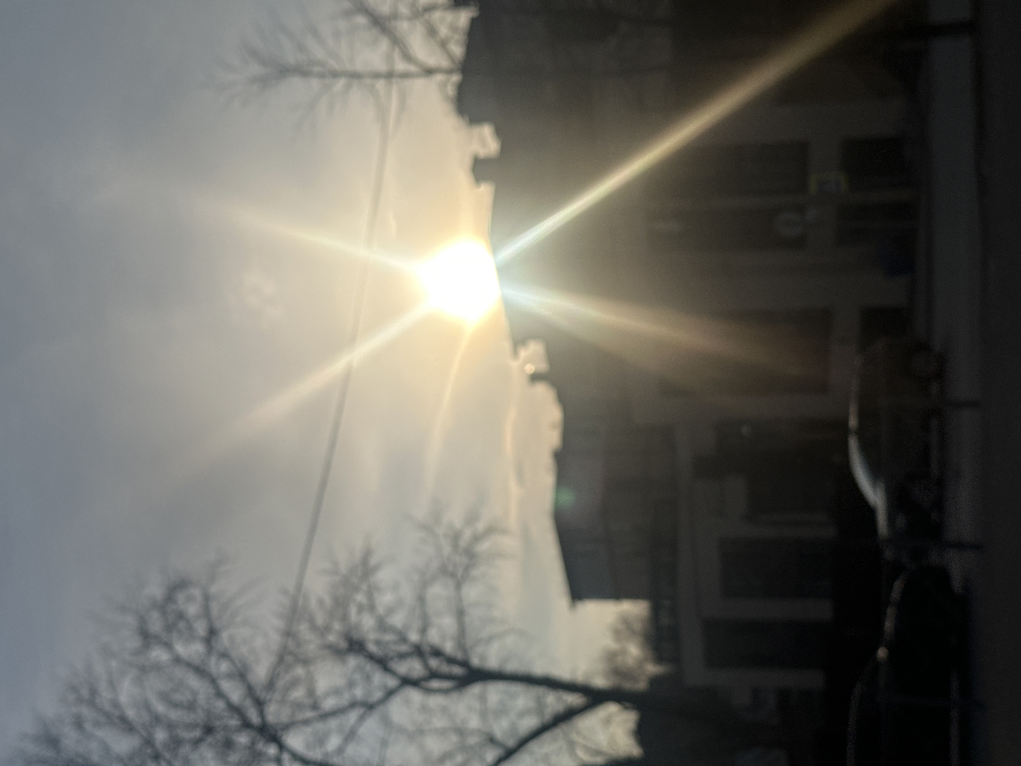 The image captures a scene in Kazan, Russia, with the sun prominently positioned in the sky, partially obscured by light clouds and the silhouette of bare tree branches in the foreground on the left. The sun's glare creates strong rays of light, partially obscuring buildings on the right side of the frame. A vehicle is parked on the right. The image appears to be taken outdoors.