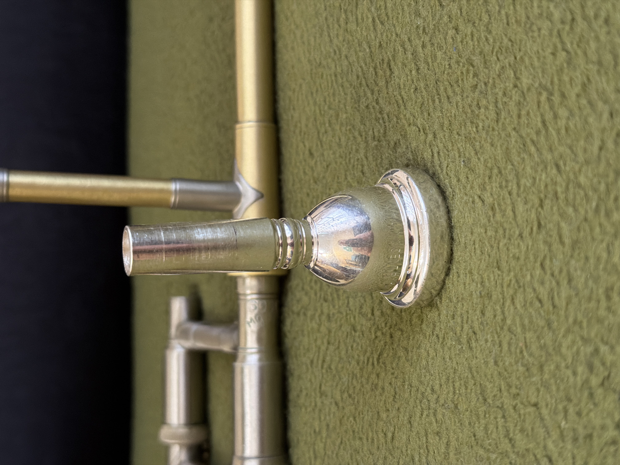 This close-up photograph captures a section of a brass wind instrument, most prominently featuring a highly reflective, silver-plated mouthpiece. The mouthpiece is inserted into the instrument's leadpipe, which transitions into a lustrous golden-brass tube. Engraved text is clearly visible on the mouthpiece, identifying it as "BACH CORP." and marked "10 1/2 C".
The instrument's components, including sections of brass and silver-colored tubing and connecting parts, are partially visible, suggesting a trombone or a similar large brass instrument. Some areas show slight wear consistent with use. The background is dominated by a richly textured, olive-green fabric, appearing soft and fibrous, potentially the lining of an instrument case or a protective surface. To the far left, a dark, out-of-focus area provides a stark contrast, possibly a shadow or another part of the environment. The lighting emphasizes the metallic sheen and intricate details of the instrument. No activity is depicted; the instrument is presented in a static, detailed view. While the context provides a location of Kyiv, Ukraine, the image itself does not contain specific visual cues to identify this location.