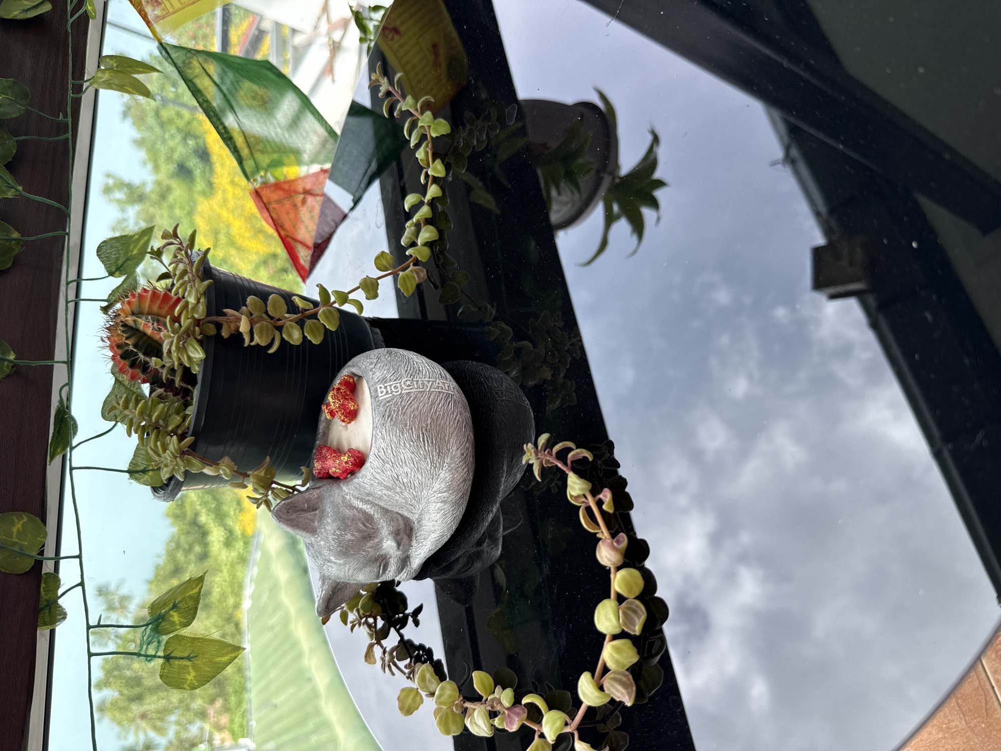 The image shows a silver, cat-shaped candle holder with two red strawberry-shaped embeds and a wick, resting on a black reflective surface. A potted cactus and succulent plant is positioned behind the candle holder. A string of prayer flags is visible in the background, along with lush green foliage and trees, suggesting an outdoor or semi-outdoor setting. The scene is likely located in a residential area or garden in the Ko Samui District of Thailand, given the tropical vegetation and prayer flags.