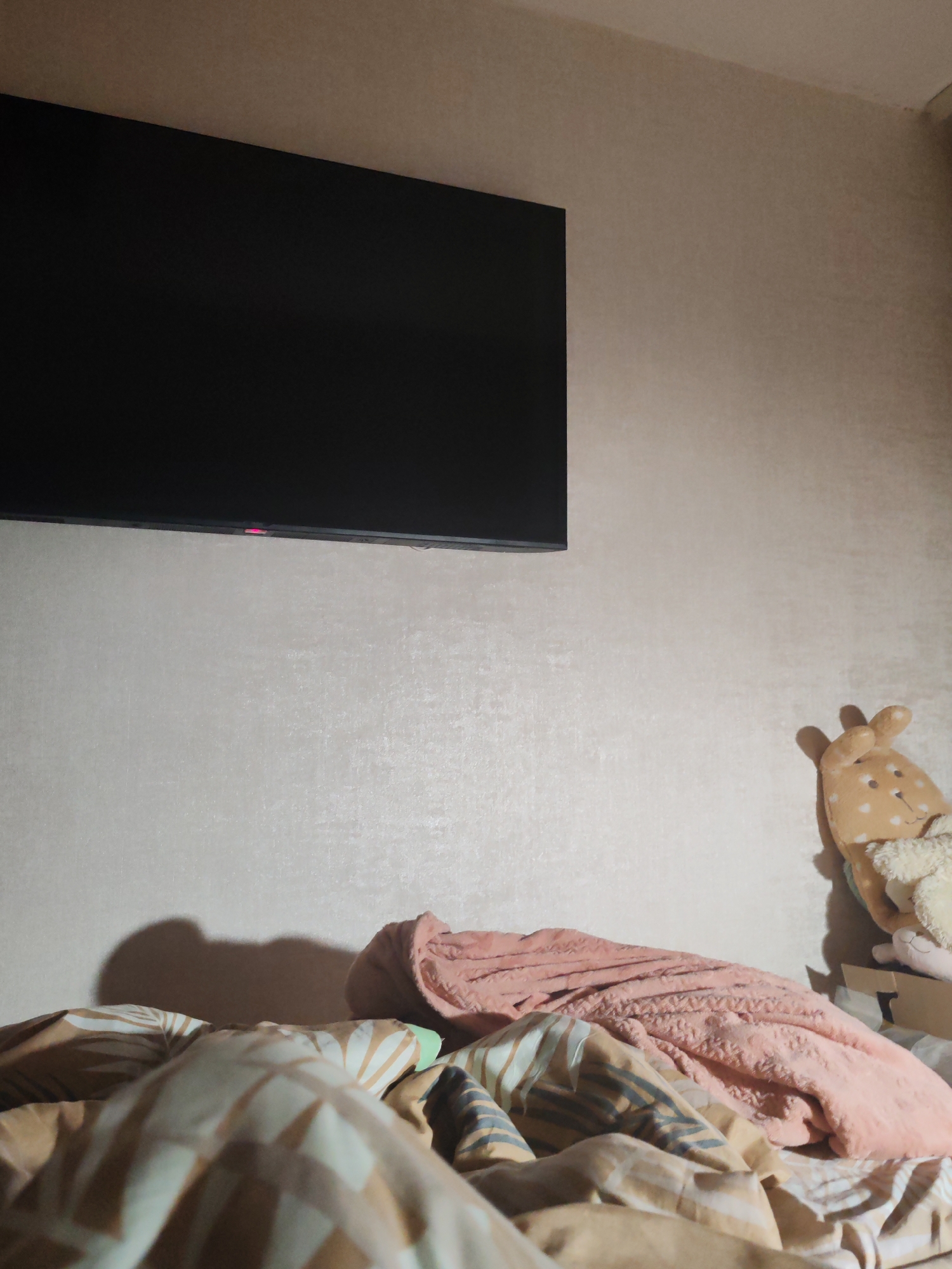 A low-angle indoor shot depicts a bed with a television mounted on the wall above it. The bed is covered with a patterned duvet and a pink blanket. A stuffed animal is propped against the wall to the right of the bed. The location is indiscernible.