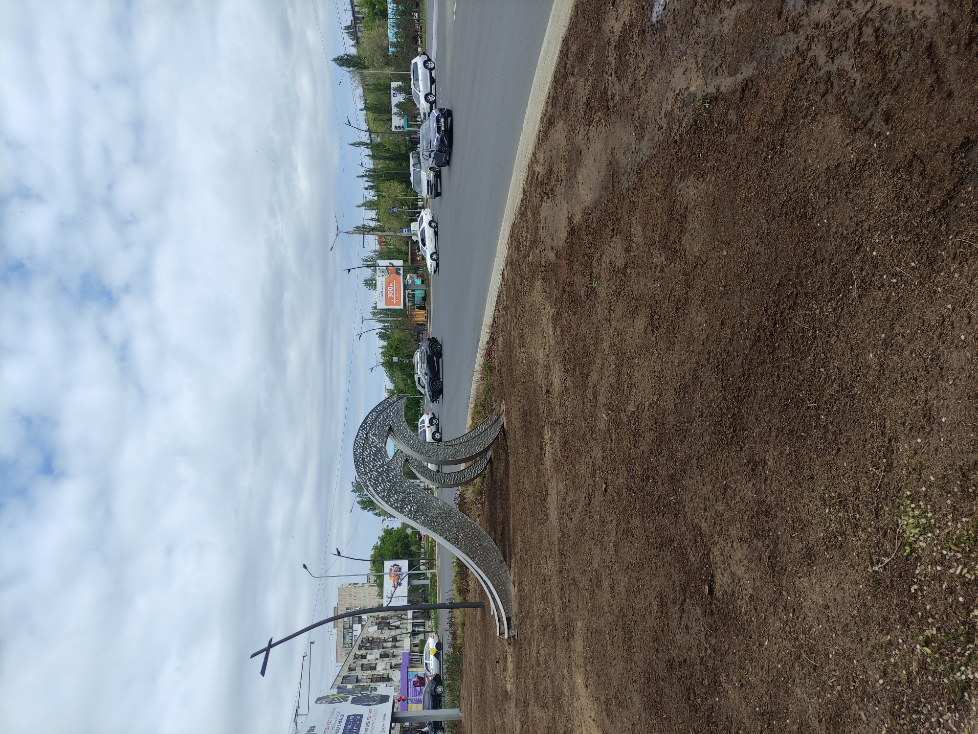 The image depicts an outdoor scene in Pavlodar, Kazakhstan. A modern, looping metal sculpture stands on a dirt patch near a road. Several cars are driving on the adjacent road. Buildings and billboards are visible in the background, and a cloudy sky is overhead. The lighting is overcast, suggesting a diffused sunlight. The location is likely a median strip between traffic lanes in a city setting.