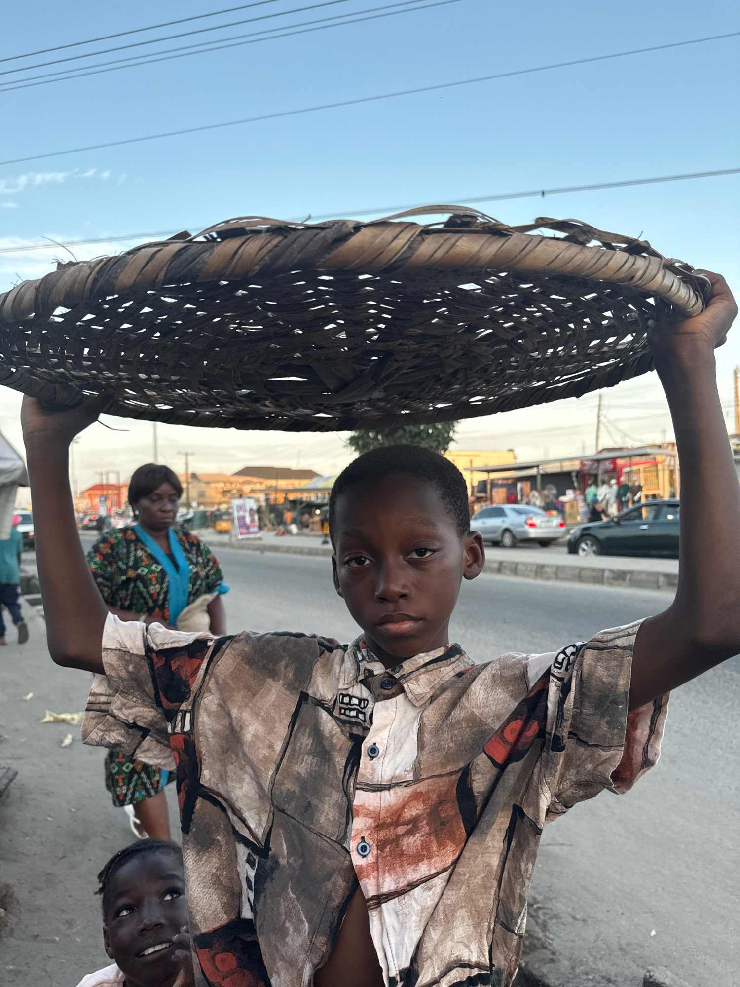 In Ado, Nigeria, a young boy, appearing to be under the age of 15, carries a large, woven basket on his head. He is dressed in a short-sleeved shirt with a busy, abstract pattern of browns, reds, and black lines. His gaze is directed forward with a neutral expression. Below and to his left, another young child, possibly a younger sibling, is visible, looking upwards with an open mouth and a smile. The background suggests an urban environment, with a paved road, several cars, and low-rise buildings characteristic of a market or commercial area. A woman in a colorful patterned dress is also visible in the middle ground, walking away from the camera. The sky above is clear with a few wisps of clouds, and power lines are visible crossing the frame.