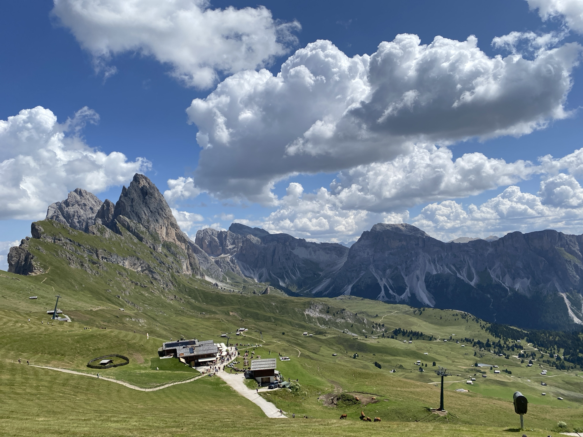 This is a wide-angle, eye-level, outdoor, daytime photograph taken in Santa Cristina Gherdëina, Italy. The image features a majestic mountain landscape with a clear blue sky filled with fluffy white clouds. In the foreground, rolling green hills are dotted with a few scattered buildings, including a rustic lodge with a wooden roof and outdoor seating. A dirt path winds through the grassy terrain, leading towards the buildings. A ski lift tower is visible on the left side of the frame, and a few cows graze peacefully in a fenced-off area in the mid-ground. In the background, towering, jagged mountain peaks rise dramatically against the sky, their rocky faces casting shadows on the verdant slopes. The overall scene depicts a serene and picturesque alpine setting, likely a popular tourist destination for hiking and outdoor activities.