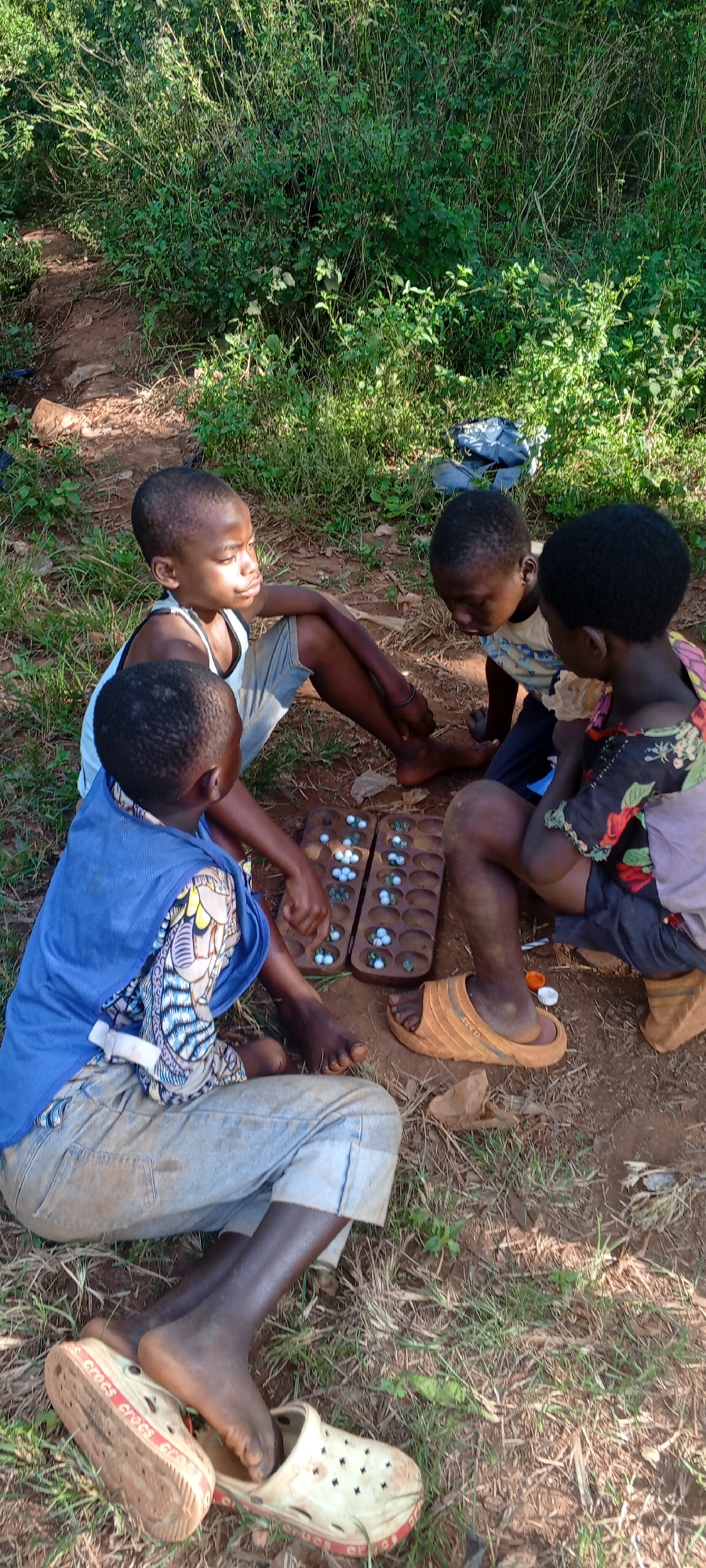 Four children are seated on the ground in an outdoor setting, possibly in a natural area or on the edge of a residential area. They are gathered around a wooden board game, which is positioned between them. The game board has rows of indentations or holes that contain white and green marbles or small stones. The children are dressed in casual clothing, including tank tops, a floral print top, and a blue vest. One child is wearing brown sandals, while another has white and red Crocs. The environment consists of dry, reddish-brown soil, patches of green grass, and some sparse vegetation. Based on the caption, this scene is set in Ngalula, Democratic Republic of the Congo.