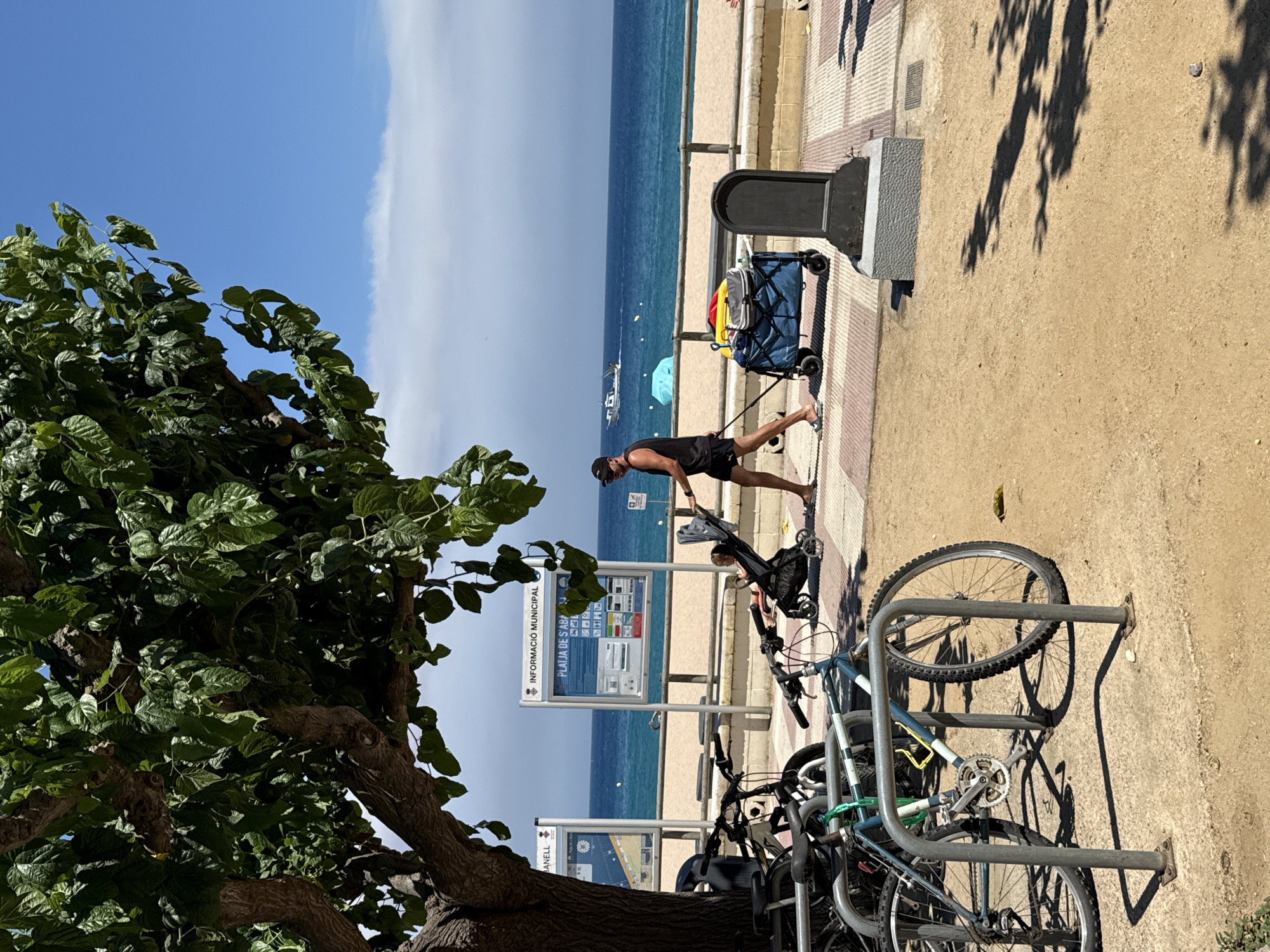 The image depicts a sunny day scene on a paved promenade adjacent to a sandy beach in Blanes, Spain. A large tree with lush green leaves shades a portion of the foreground. In the immediate foreground, several bicycles are parked in a rack. To the right of the bikes, a man wearing a black tank top, black shorts, and a black baseball cap is pulling a blue wagon loaded with yellow and gray items. He is walking on the promenade. Behind him, a stroller containing a child is positioned near a black monument. Further back, the sandy beach stretches to meet the blue ocean, with a few small boats visible in the distance. Informative municipal signs are posted along the promenade. The sky is bright blue with a few wispy white clouds.