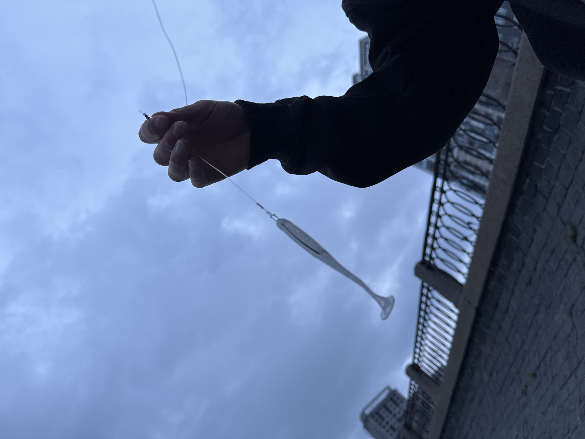 A person in a black hooded sweatshirt holds a fishing line with a silver lure attached. The person's left hand is visible, gripping the line near a small weight. The lure, shaped like a small fish, dangles from the line. The scene is set against a cloudy, grey sky. In the background, a decorative metal railing lines a pathway. Beyond the railing, buildings are visible, some appearing to be of modern architecture. The ground in the foreground is paved with cobblestones. This setting suggests a location along one of the many embankments in Saint Petersburg, Russia, likely near the Neva River or one of its canals.