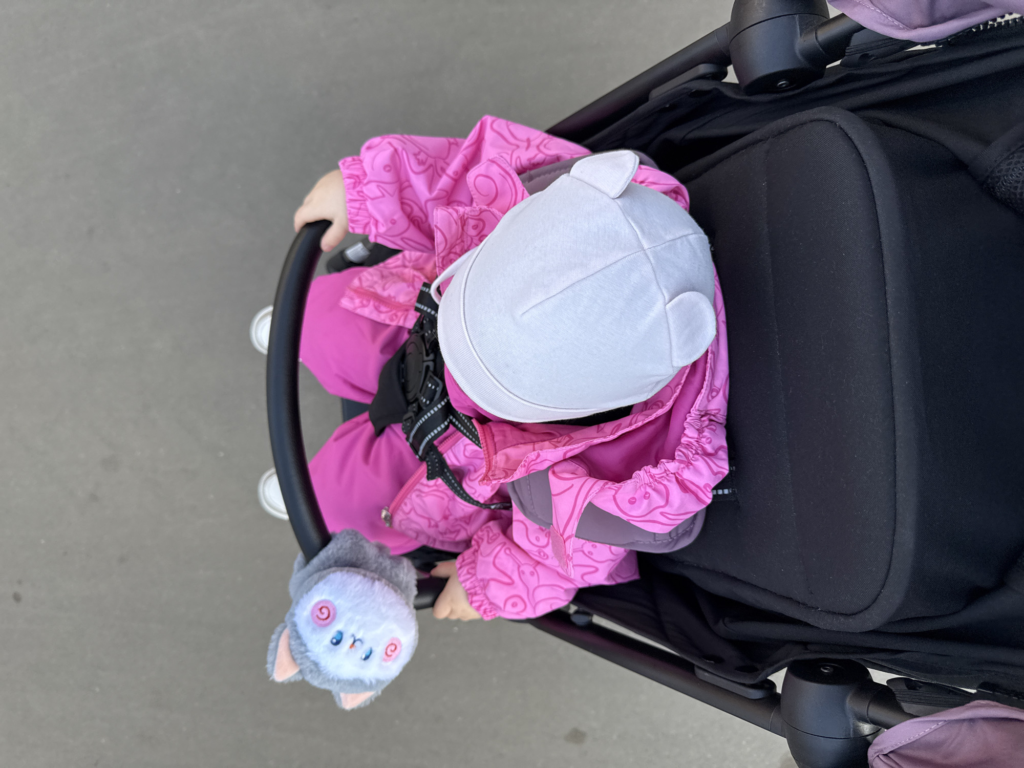 A high-angle shot shows a young child sitting in a stroller. The child is wearing a bright pink snowsuit and a white cap with small bear ears. A small plush owl toy is attached to the stroller's handle. The stroller is black, and the seat is facing away from the camera. The setting is a grey paved surface, likely a sidewalk. The image does not contain enough information to determine the location in Moscow, Russia.