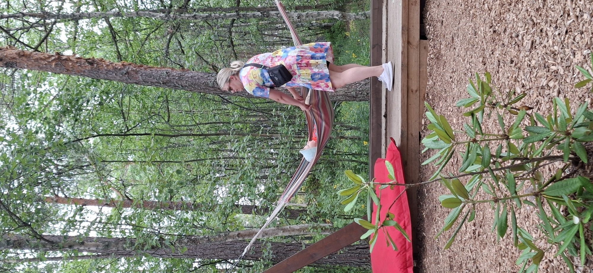 The image depicts a woman standing on a wooden platform, holding onto a hammock. A baby is lying in the hammock, wearing a blue bonnet and a red garment. The woman is wearing a colorful floral dress and a black crossbody bag. The hammock is striped with various colors. Behind them, a dense forest with tall trees and green foliage is visible. In the foreground, a small green plant with broad leaves grows out of a bed of wood chips. A large red cushion is placed on the platform to the left of the hammock. The scene appears to be an outdoor recreational area, possibly part of a park or a resort in Naantali, Finland. The lighting suggests it is daytime, with overcast conditions.