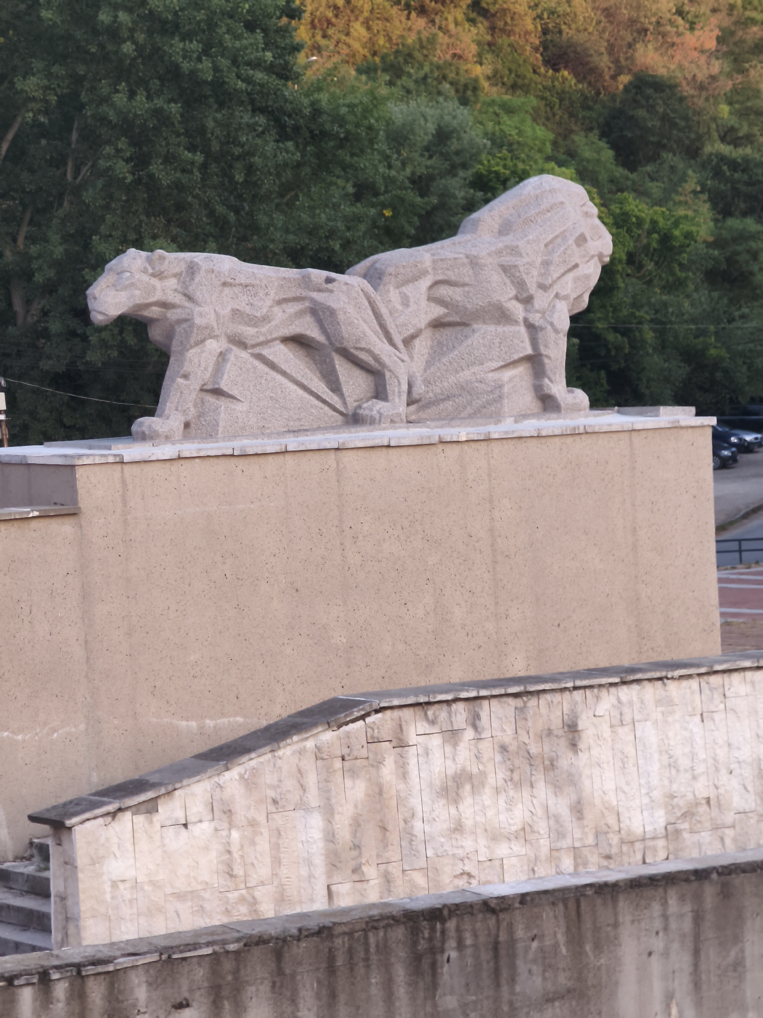 The image shows a stone sculpture of two lions positioned atop a building in Lovech, Bulgaria. The lions are carved in a stylized, angular manner, with the one on the left facing left and the one on the right facing right. They appear to be resting or guarding. The building beneath them is constructed of a tan, textured material, with a lighter stone facade visible on a lower level, featuring a staircase. In the background, dense green trees are visible, with some areas of autumnal foliage and hints of sunlight. A few cars are parked on a road in the distance. The overall scene depicts an outdoor monument, likely a landmark in the city.