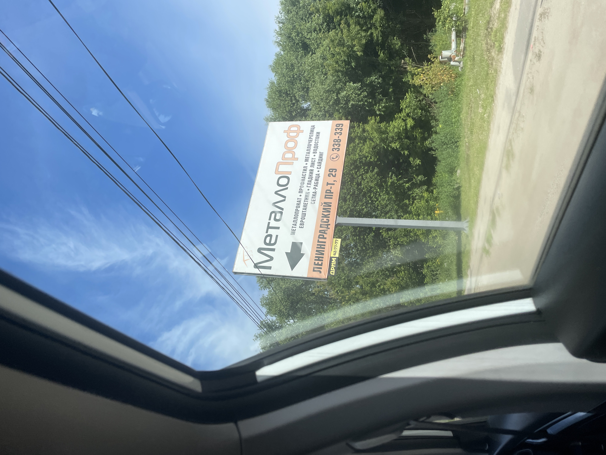 The image shows a billboard with the text "МеталлоПроф" in Russian, meaning "MetalProff". The billboard also lists various metal products such as "металлопрокат", "профнастил", "металлочерепица", "евротантагник", "гладкий лист", "водостоки", "сетка-рабица", and "сайдинг". Below this, it indicates "ЛЕНИНГРАДСКИЙ ПР-Т, 29" which translates to "Leningradsky Prospekt, 29" and a phone number "338-339". There is also a smaller text "дпм 42-77-77" which might be another contact or identifier. The billboard is positioned outdoors, with trees and a blue sky with clouds in the background. The perspective suggests the photo was taken from a moving vehicle, looking out of a window. The scene is identified as being in Rusanovo, Russia.