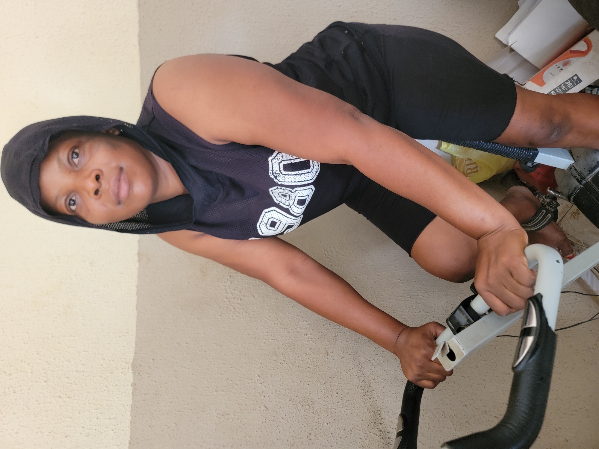 The image, taken in Sabon Tasha, Nigeria, captures a person engaging with an exercise bike in an indoor setting. The individual, appearing to be female with dark skin, is shown from the chest up, leaning forward on a stationary bicycle and holding its handlebars with both hands. She is wearing a black sleeveless hooded top that features white outlined text, where "BO" is clearly visible, and black shorts. A distinct ankle bracelet adorns her right ankle.
Her head is slightly turned towards the camera, and she maintains a calm, neutral expression with a soft gaze. The background features a light-colored, textured wall. To her right, behind the exercise equipment, various items are visible, including cardboard boxes, a yellow plastic bag, and a white and orange appliance. This appliance has a digital display showing numbers like "36", "18.3", "23%", and "58kg," suggesting it might be a body composition scale or a fitness tracker. The ambient lighting indicates it is daytime. The scene portrays a moment of personal fitness activity within a casual, likely home-based, environment.