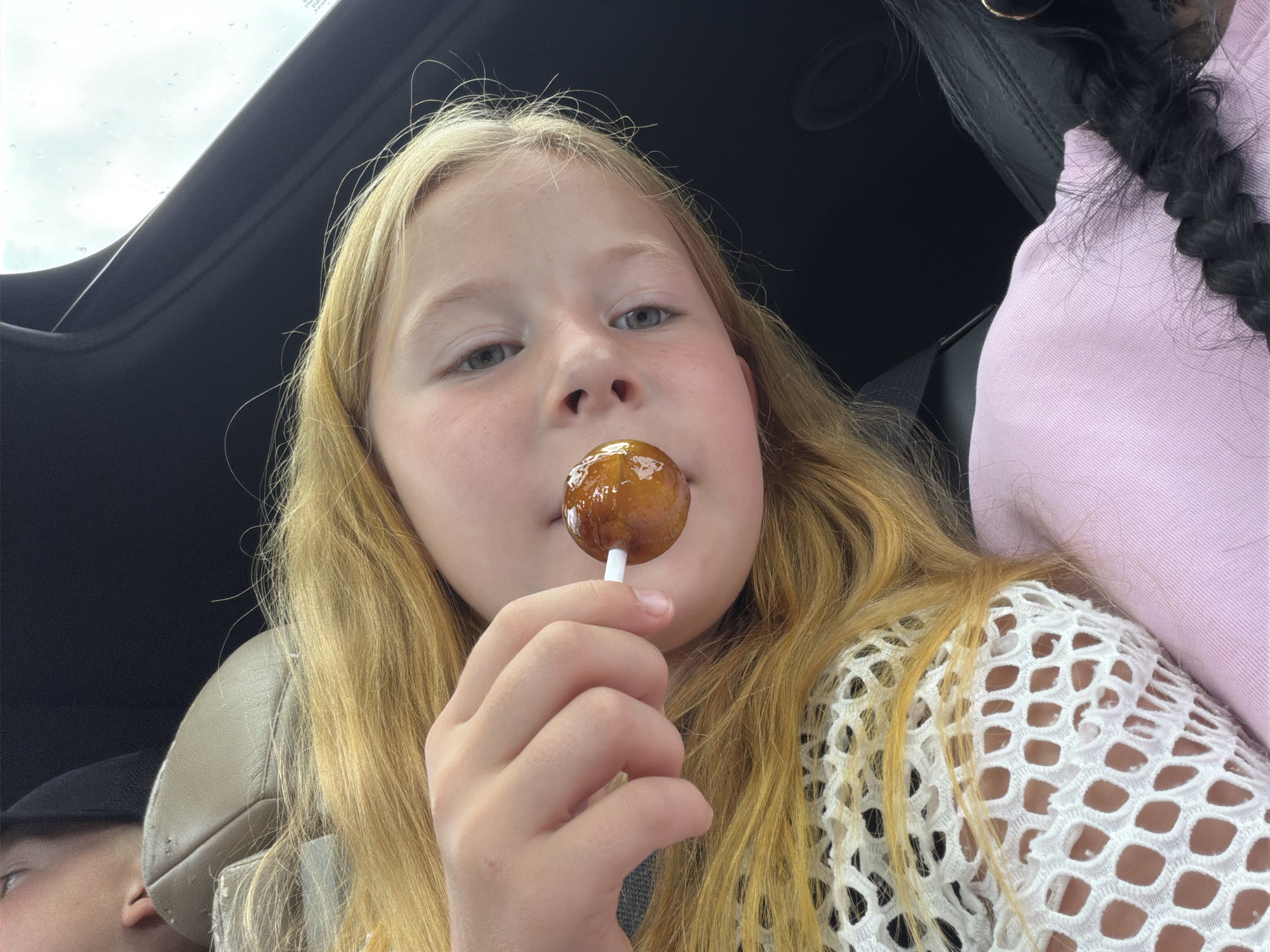 A young girl with blonde hair is holding a lollipop to her mouth. She is in a car, with a view of the sky through the sunroof. The sky appears overcast. Another person with dark hair in a braid, wearing a pink top, is visible in the background. The girl is wearing a white, mesh-like garment. The scene suggests a car ride, possibly on a cloudy day in Шолохово, Russia.