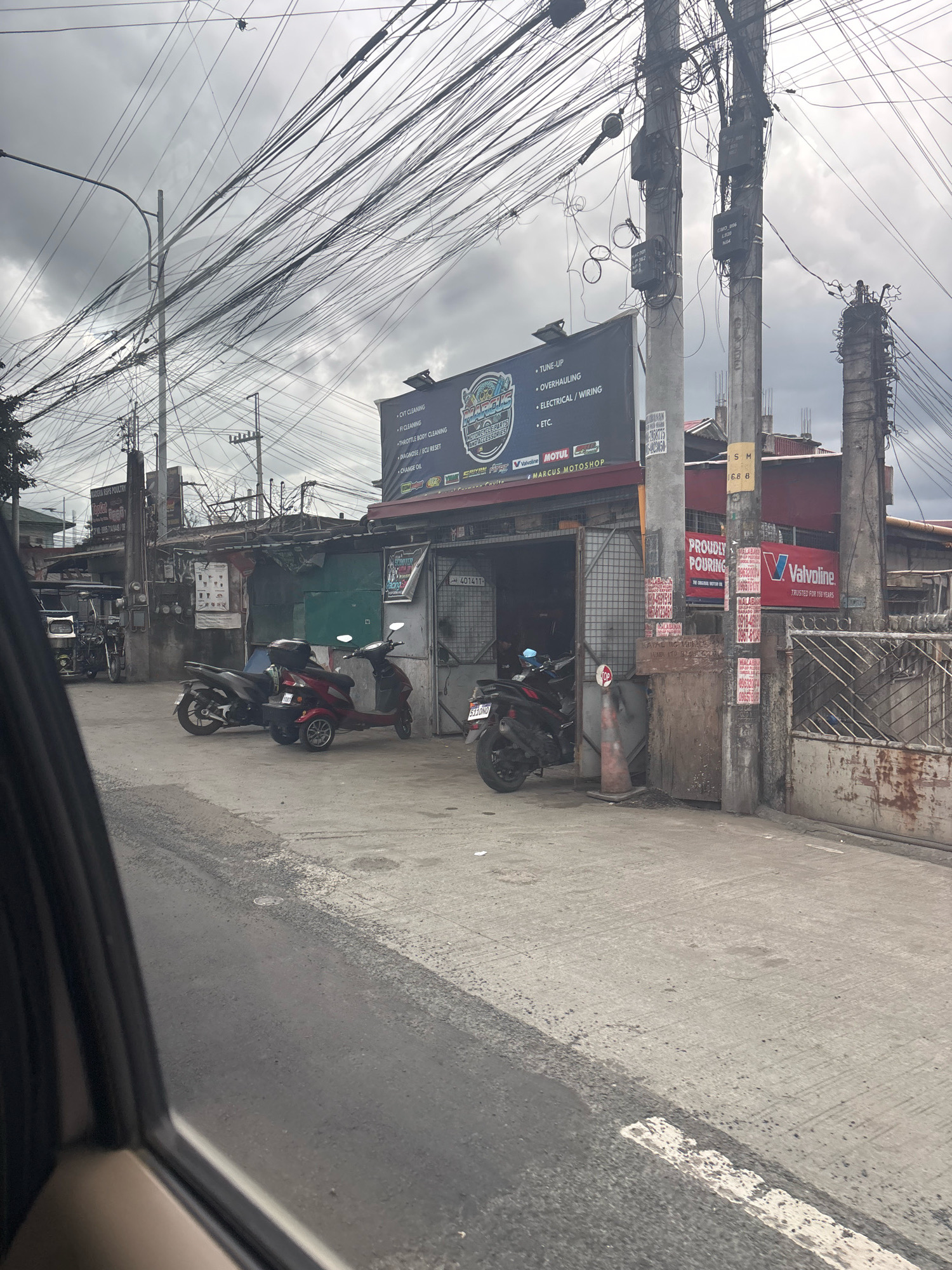 A street-level, medium shot captures a street scene in Carmona, Philippines, dominated by a tangled web of overhead electrical wires and utility poles. The buildings visible are low-rise structures, some appearing to be shops or service centers.
In the foreground, the asphalt road shows signs of wear and tear. On the right side of the road, a gate made of corrugated metal and diamond-patterned bars is partially visible. Attached to a utility pole near the gate is a red and white Valvoline sign. Several flyers and posters are adhered to the utility pole and the adjacent wall.
A partially open garage-like structure is situated on the left side of the image, with a green metal door visible. Two motorcycles are parked in front of the establishment. The motorcycle on the left is dark gray, and the one on the right is a red scooter with a sidecar-like modification featuring a third small wheel. A black motorcycle with a red accent is parked further inside the garage.
A large, illuminated billboard is mounted above the garage, displaying text and logos related to motorcycle services. Some of the visible text includes "TUNE-UP", "OVERHAULING", "ELECTRICAL/WIRING", "FI CLEANING", "THROTTLE BODY CLEANING", "DIAGNOSE/ECU RESET", "CHANGE OIL", and "ETCS". The name "MARCUS MOTOSHOP" is prominently displayed on the billboard.
Further to the left, another structure is visible with a smaller sign that reads "ROSA POULTRY". In the far left of the frame, a tricycle-like vehicle is parked.
The sky is overcast, with heavy clouds suggesting impending rain or a generally gloomy atmosphere. The overall impression is of a typical urban or semi-urban street in the Philippines, characterized by its dense electrical infrastructure and commercial activity.