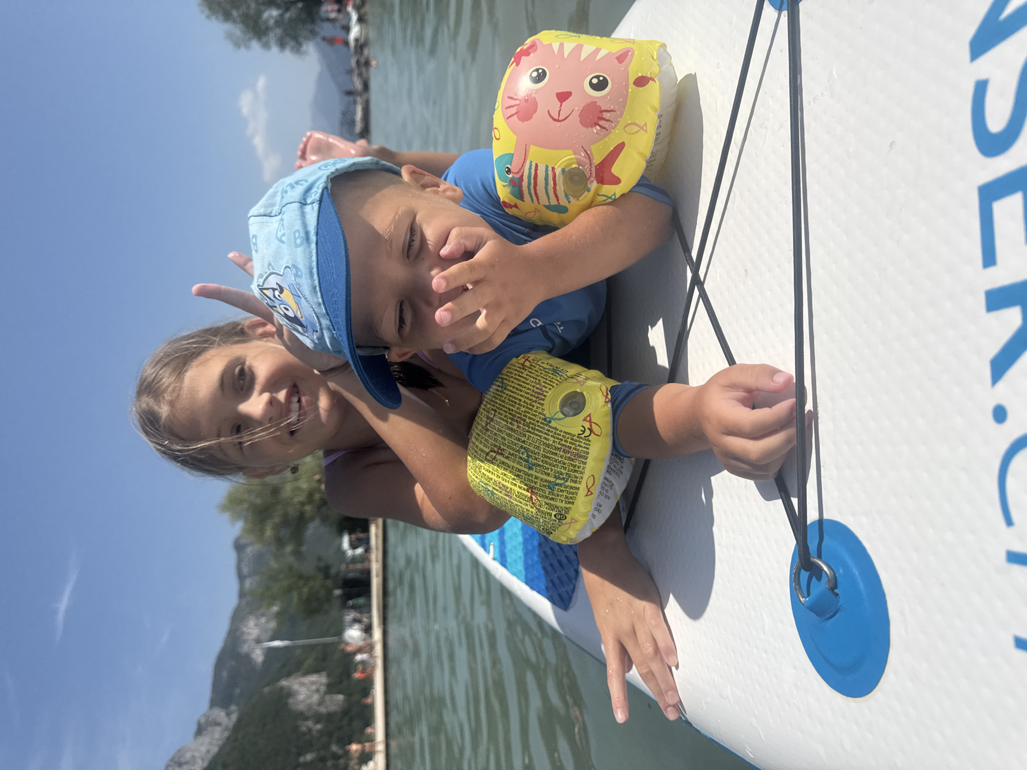 Two children, a girl and a boy, are lying on a white paddleboard on the surface of Lake Annecy. The girl, positioned behind the boy, is smiling and making a peace sign with her fingers. She is wearing a pink swimsuit. The boy, in the foreground, is wearing a blue t-shirt and a blue baseball cap with a cartoon character on it. He has yellow inflatable armbands on, one with a cartoon pig and the other with text. The boy is covering his mouth with his hand and his eyes are closed. The paddleboard is equipped with a blue loop and bungee cords. The background shows the clear blue sky, a body of water, and mountains. The scene appears to be a sunny day on the lake.
