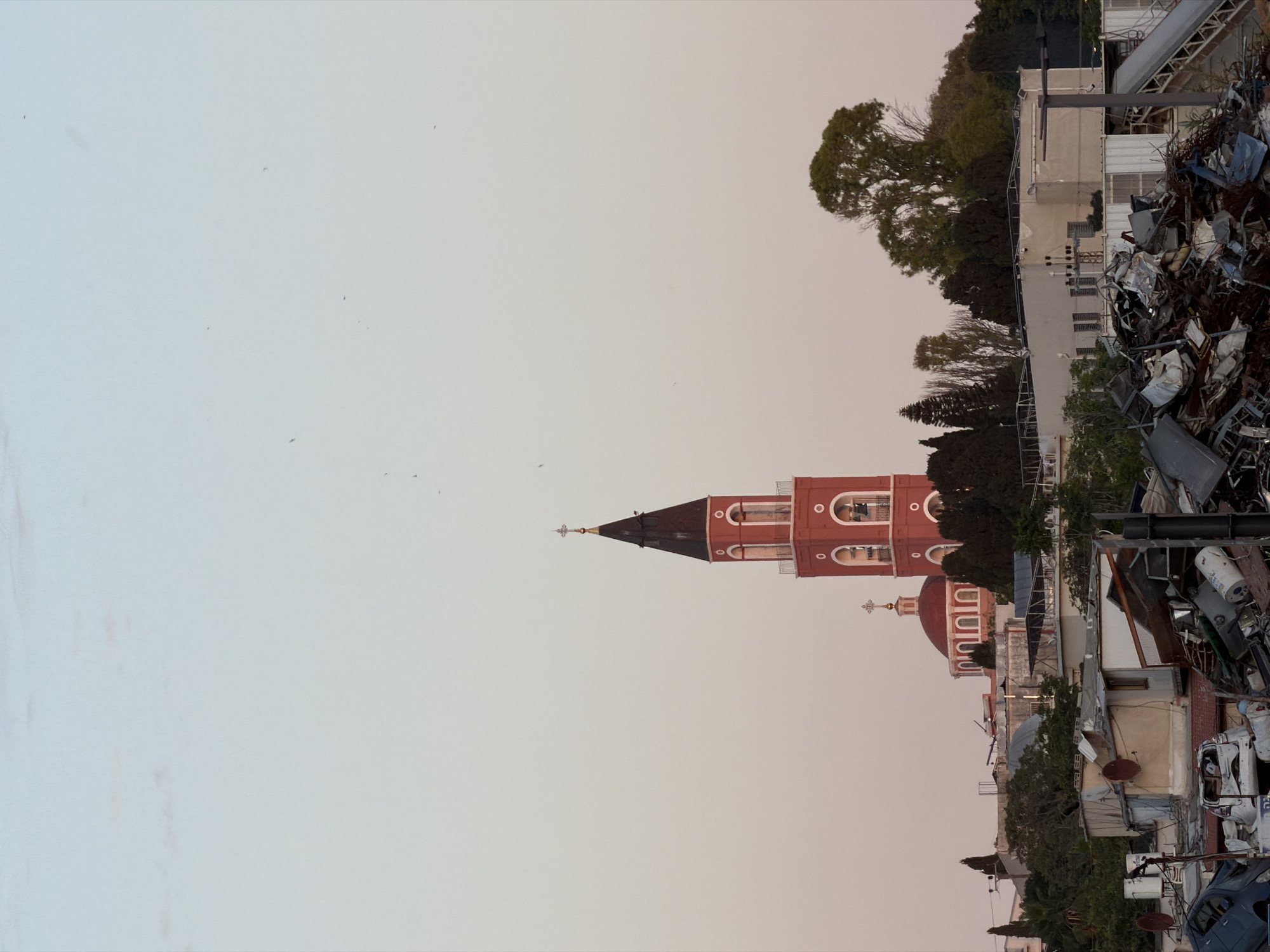 The image shows a view of Tel Aviv, Israel. In the center, there is a red church with a tall steeple and rounded domes. The sky above is light and hazy. Clutter and discarded objects, including what appears to be scrap metal and parts of vehicles, are in the foreground. Buildings and greenery are adjacent to the church.