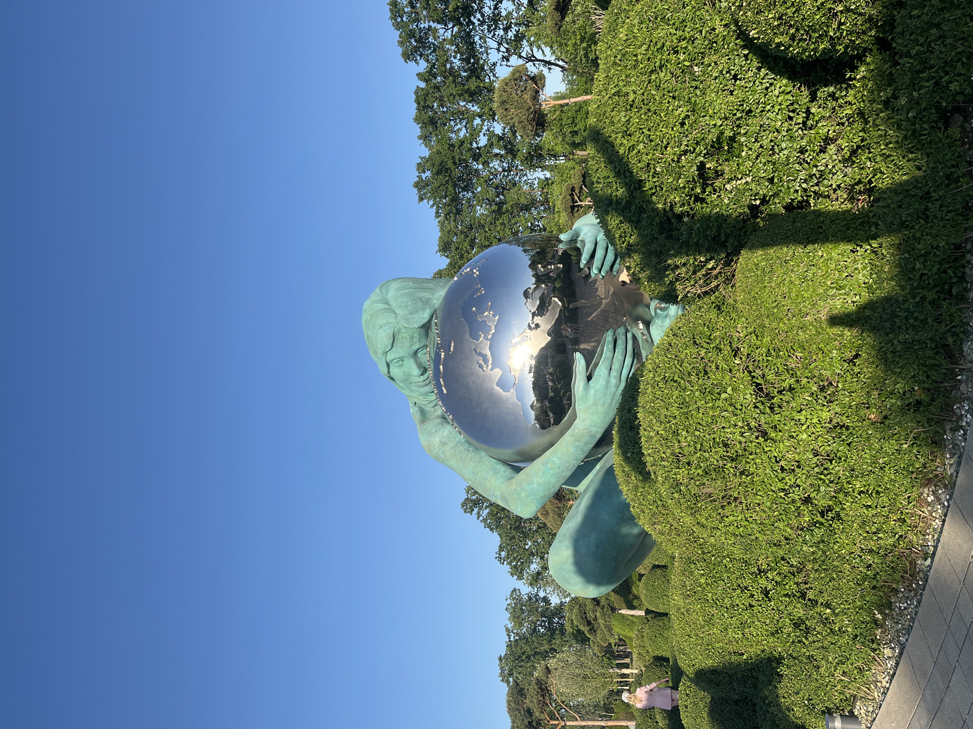 The image shows a sculpture of a woman holding a reflective globe. The sculpture is located in a park, surrounded by manicured bushes and trees. The sky is clear blue. Based on the presence of the "Unity of the World" sculpture by Oleg Ershov, the location is most likely the Galitsky Park in Krasnodar, Russia.