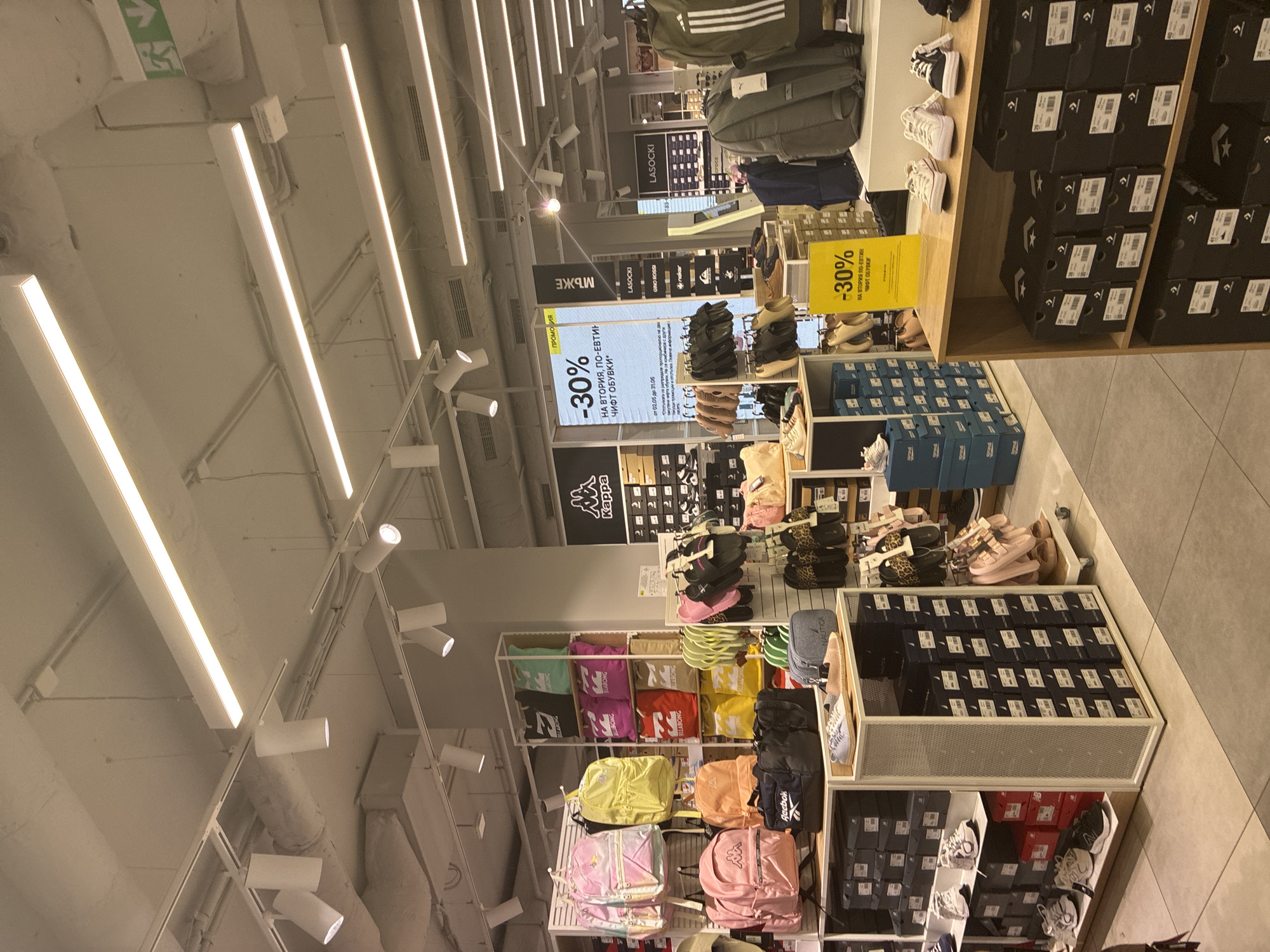 The image captures the interior of a retail store. Fluorescent strip lights illuminate rows of shelves and display units stocked with merchandise, including backpacks, shoes, and shoeboxes. Signage advertises a "30% off" promotion on a second, less expensive pair of shoes. The store appears to be located in Plovdiv, Bulgaria, as signage written in Cyrillic includes the word "ОБУВКИ" (shoes), and the brands Kappa, Reebok, and Gino Rossi. There are no individuals present in the frame.